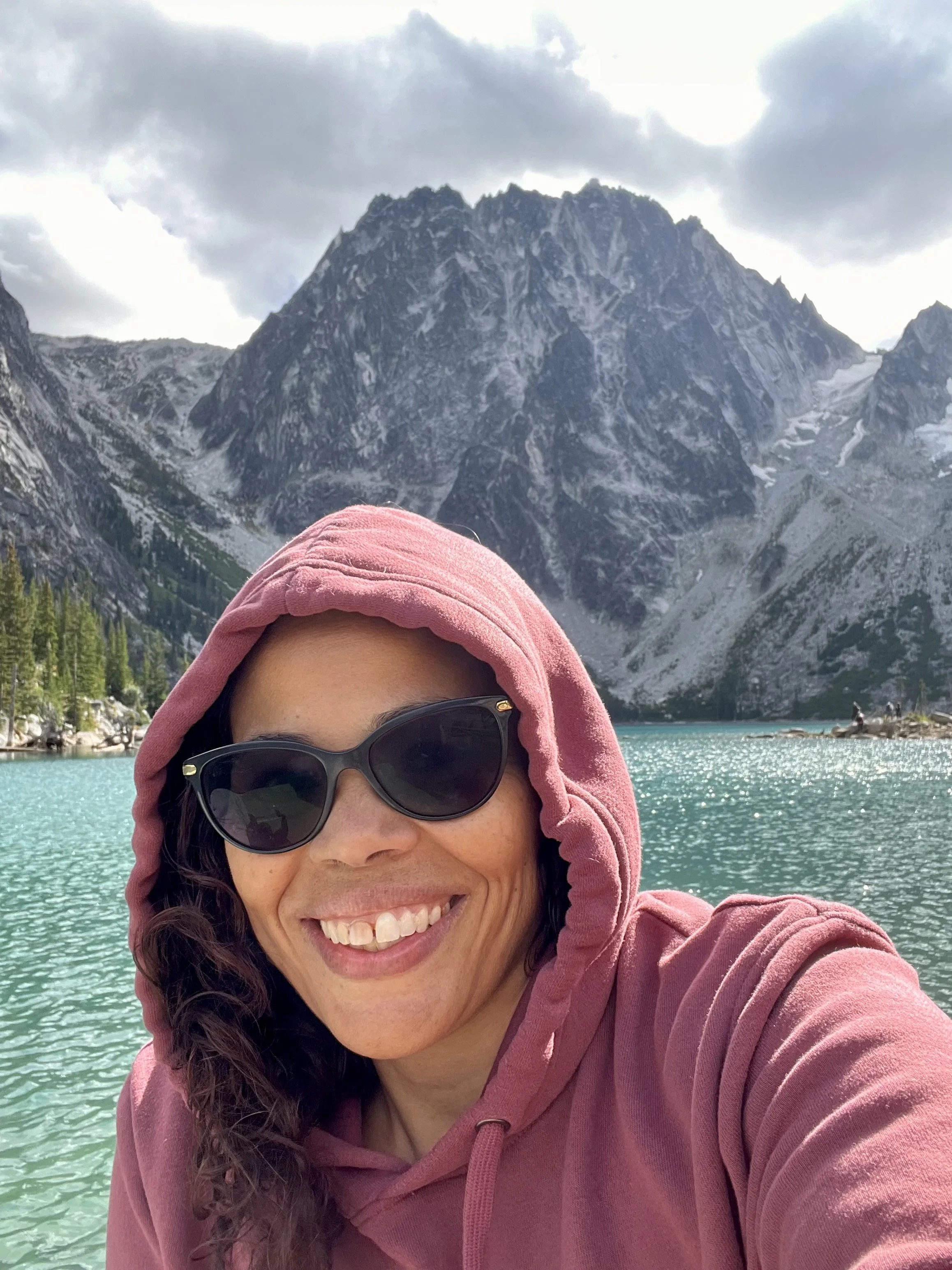 Close-up of a woman smiling, wearing sunglasses and a pink hoodie, with a mountain lake in the background.