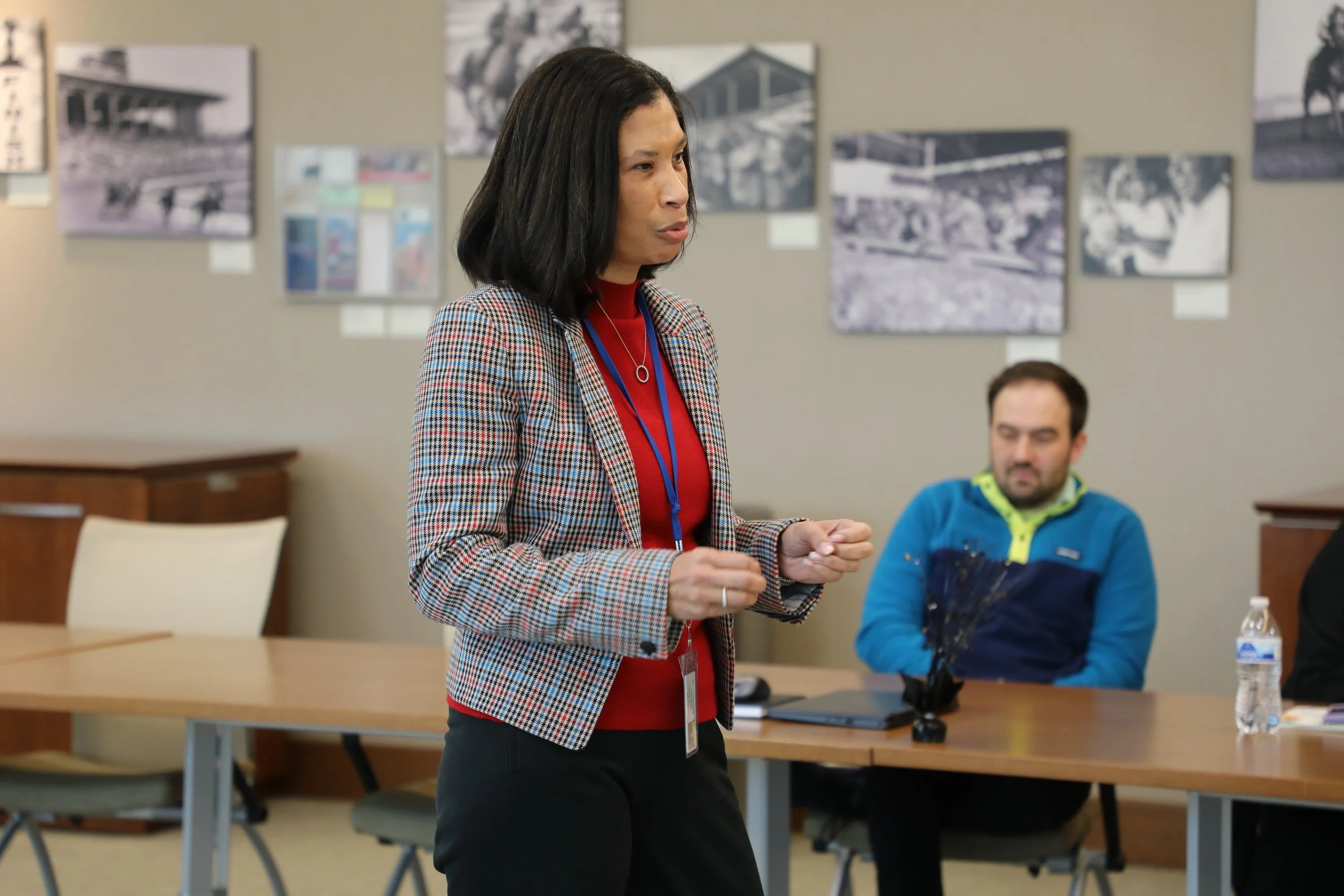 A woman giving a presentation in a conference room with a man sitting at the table behind her. The background has framed black and white photos on the wall.