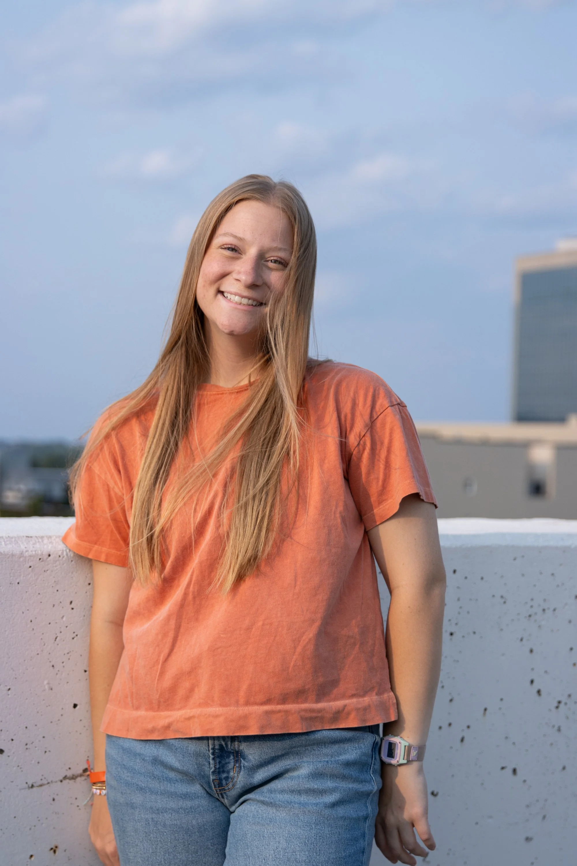 A young woman with long red hair wearing an orange T-shirt and blue jeans, smiling on a rooftop with a cityscape in the background.