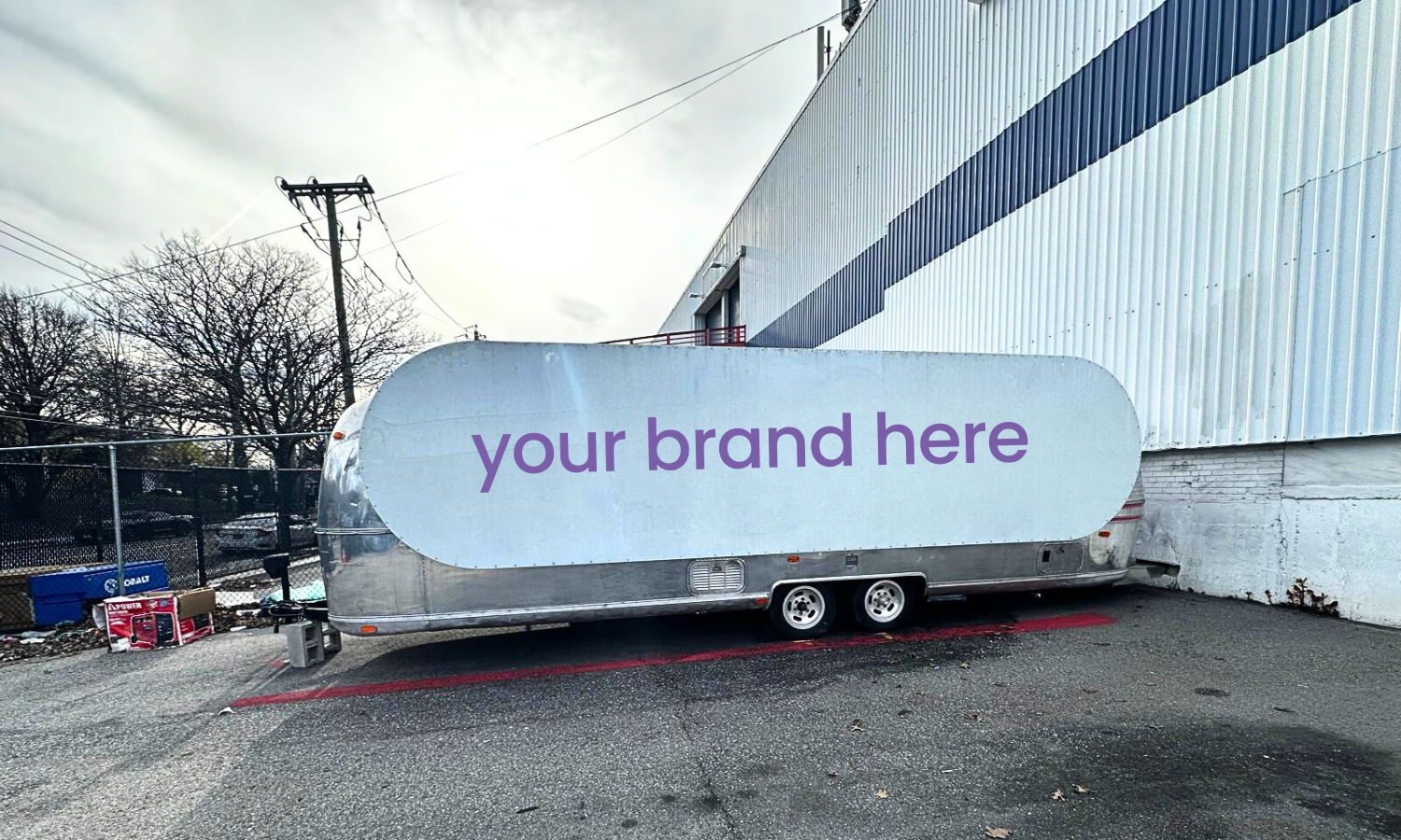 A shiny, silver, vintage travel trailer with a large sign on the side that reads 'your brand here' in purple text. The trailer is parked on an asphalt lot next to a white industrial building with blue siding, with some trees and utility poles in the background.