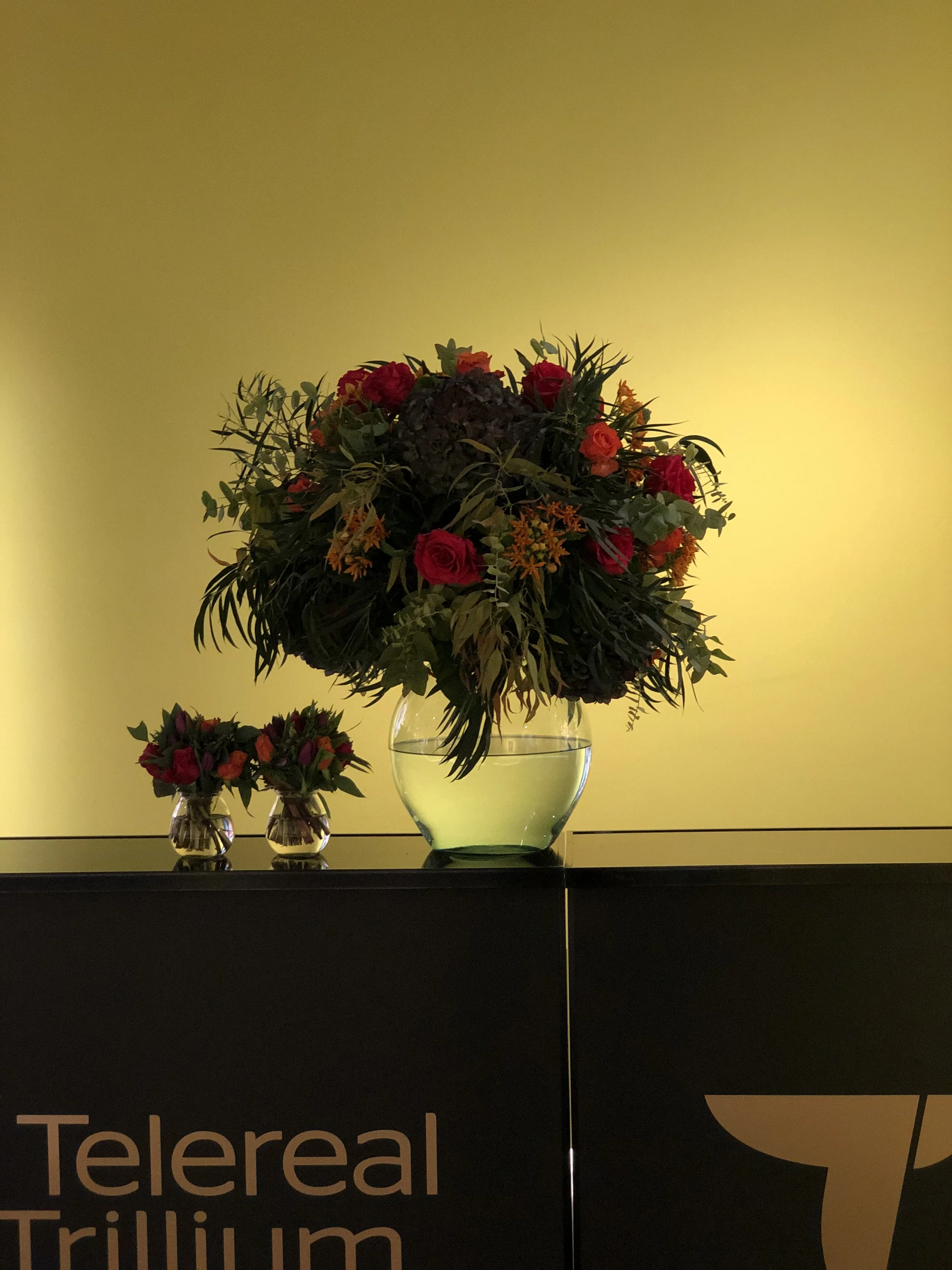 Large floral arrangement in a rounded glass vase with water, placed on a dark surface against a yellow background, with two smaller matching floral arrangements in tiny vases beside it.