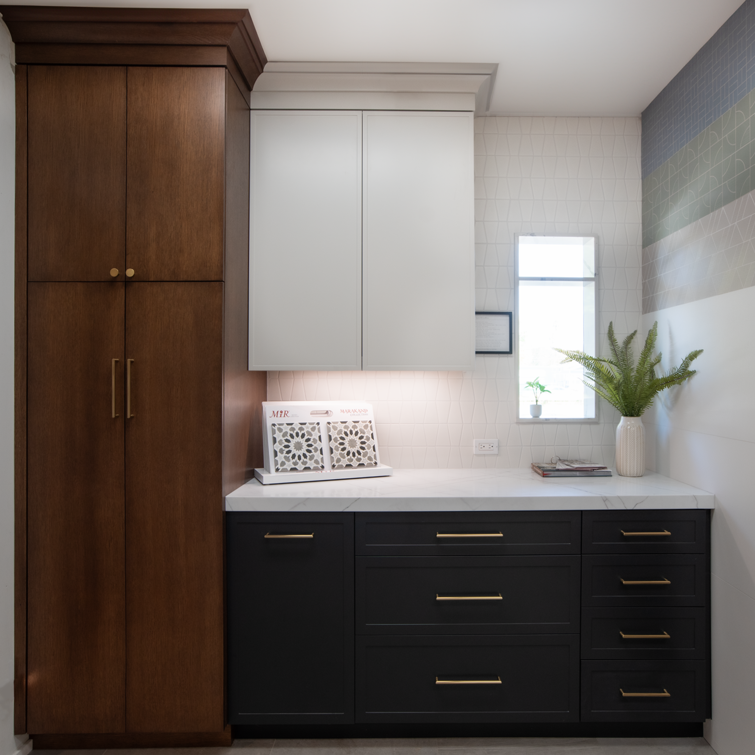 Interior of a modern kitchen workspace with black and white cabinets, a white marble countertop, a small window, and decorative plants.