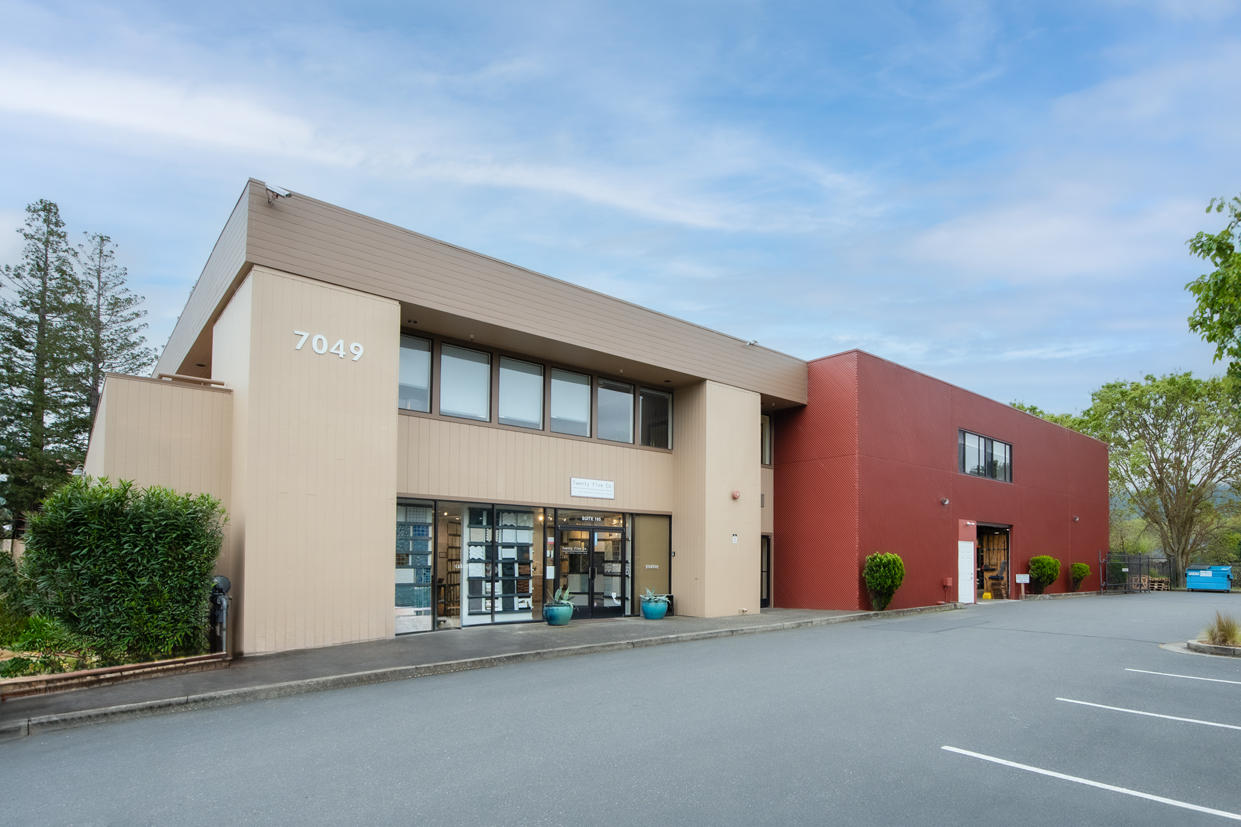 Modern two-story commercial building with beige and red exterior walls, large windows, parking lot in front, and trees in the background.