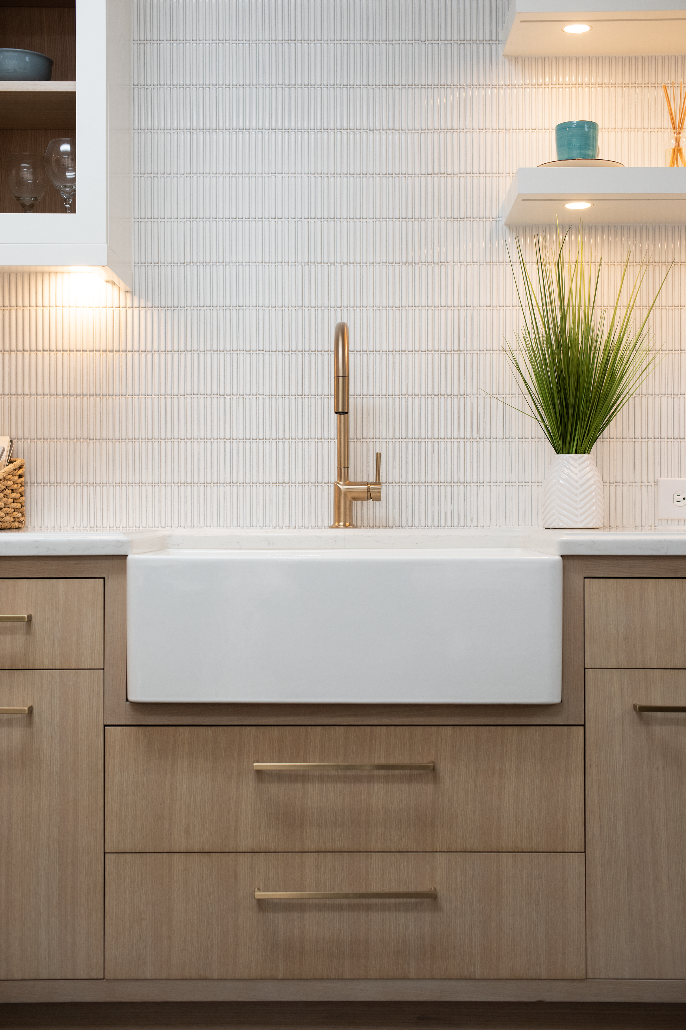 Modern kitchen with a farmhouse sink, beige cabinetry, a gold faucet, a potted plant, and open shelves with decor items, illuminated by recessed lighting.