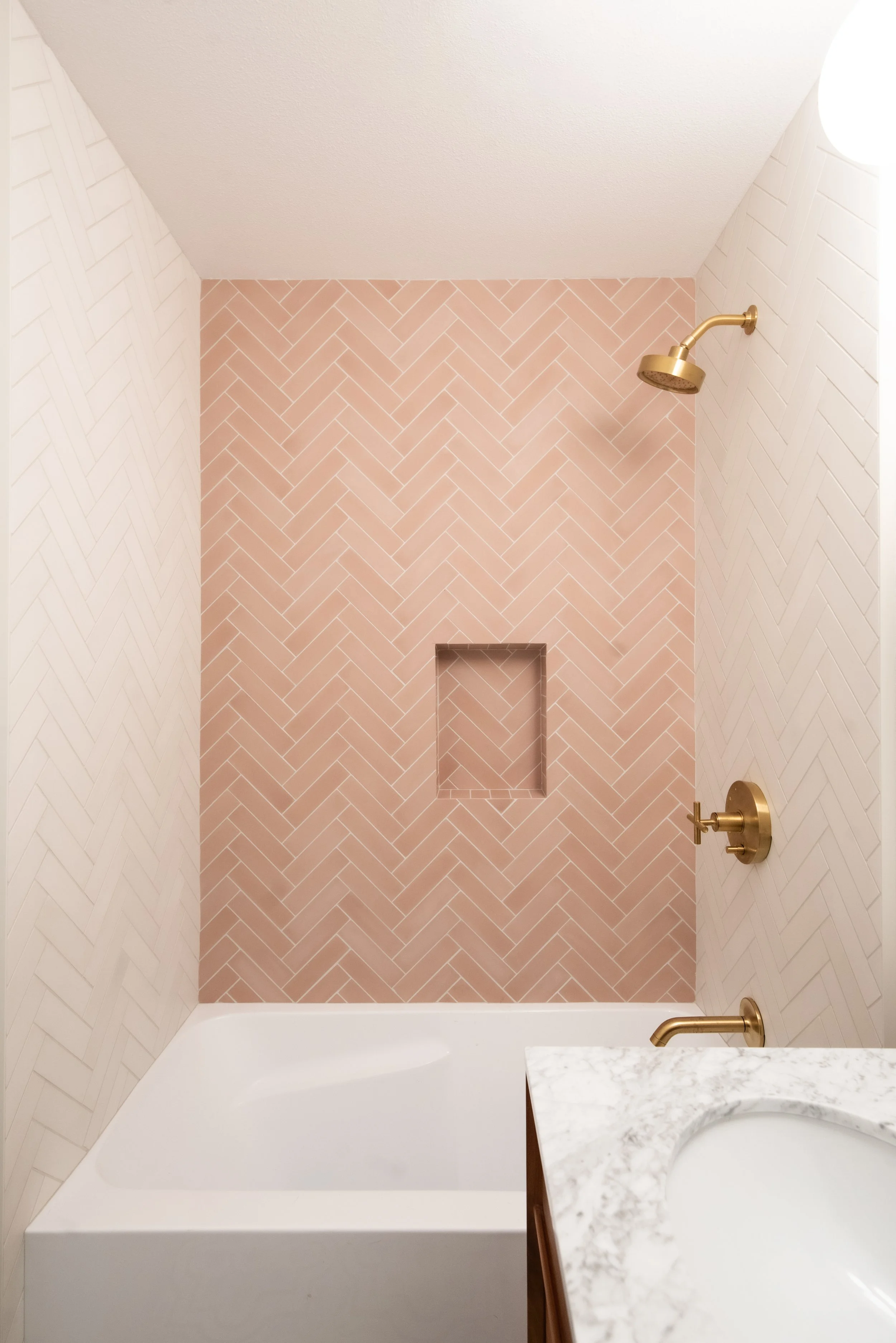 A bathtub and a marble countertop with a sink in a bathroom with a pink herringbone tile wall and gold fixtures.