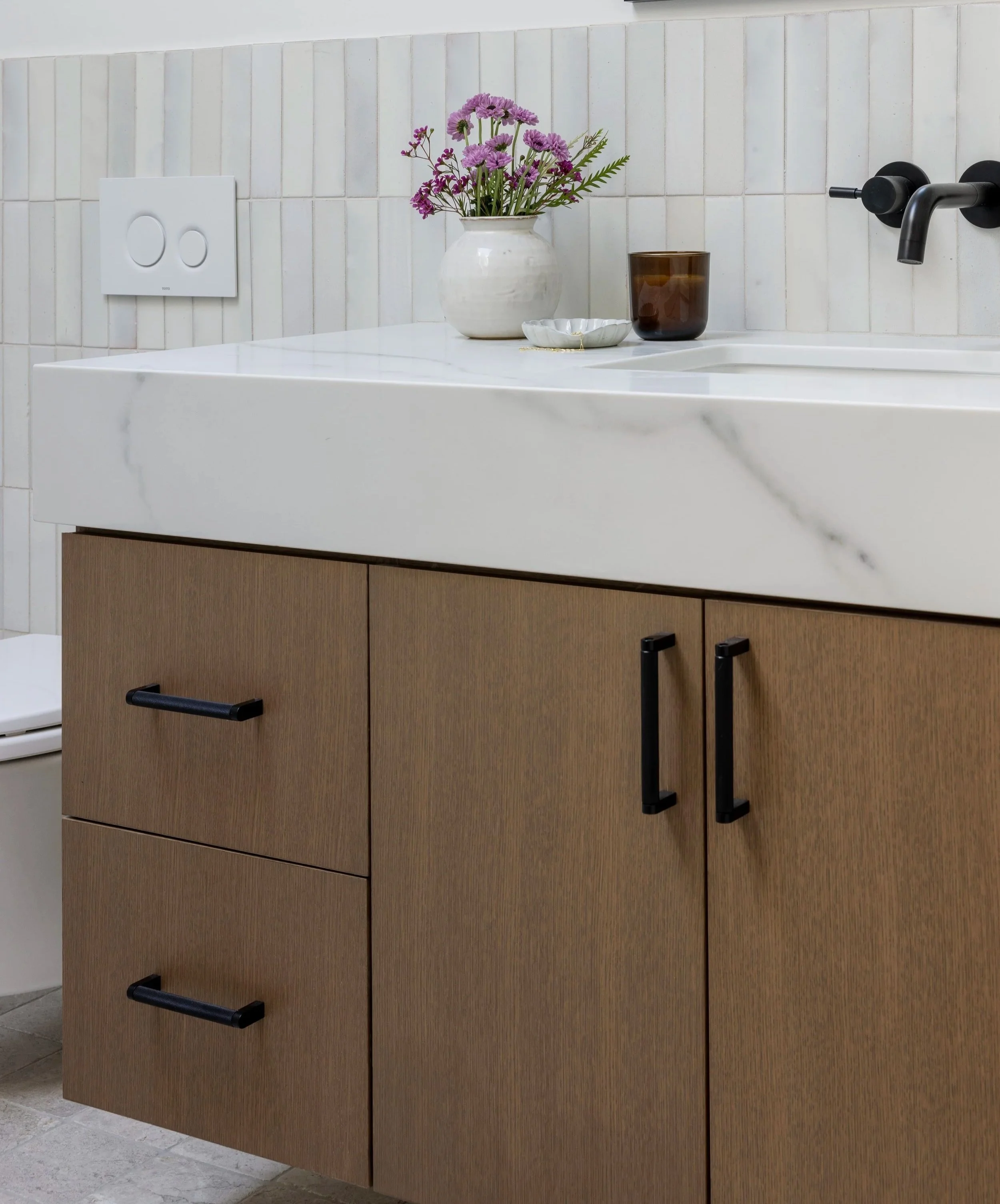 Modern bathroom vanity with a white marble countertop, black handles on wooden cabinet doors, a white vase with purple flowers, a brown glass, and a white dish.