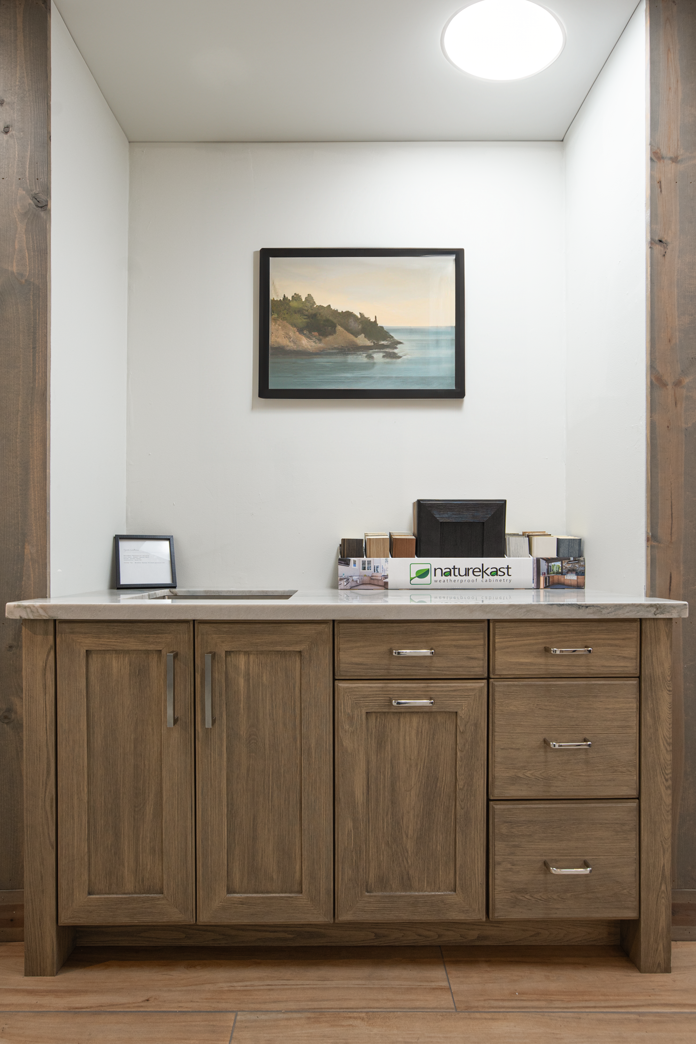 Interior view of a wooden cabinet with a marble countertop in a showroom or office, featuring a framed landscape painting of a coastline, a small framed document, and a display box with samples and a black frame on top.