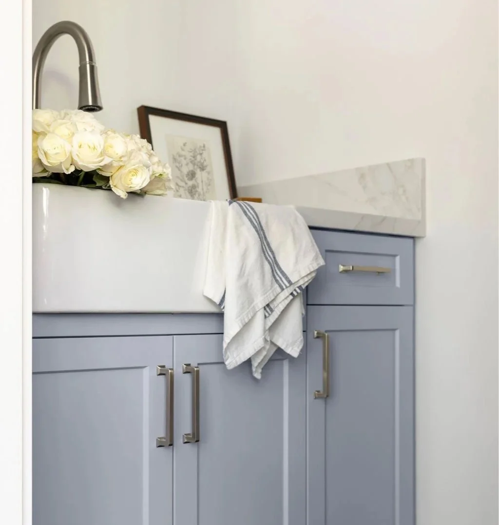 Light blue bathroom cabinet with gold handles, a white countertop with a marble backsplash, a towel hanging over the edge, a framed botanical print leaning against the wall, and a bouquet of white roses near a kitchen faucet.