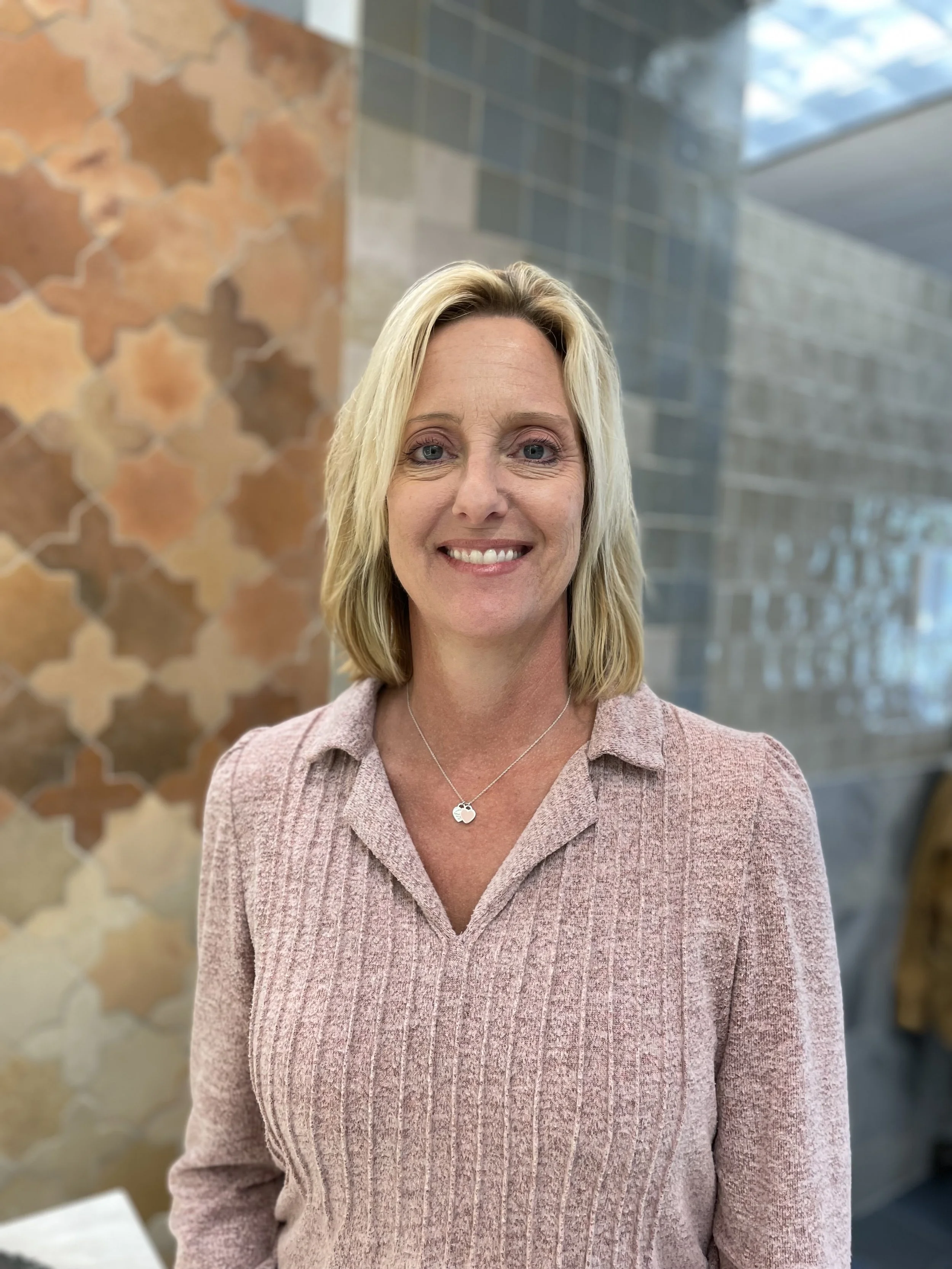A woman with blonde hair, smiling, wearing a pink ribbed sweater and a silver necklace with two silver circles, standing indoors in front of a decorative wall with brown tiles on the left and gray tiles on the right.