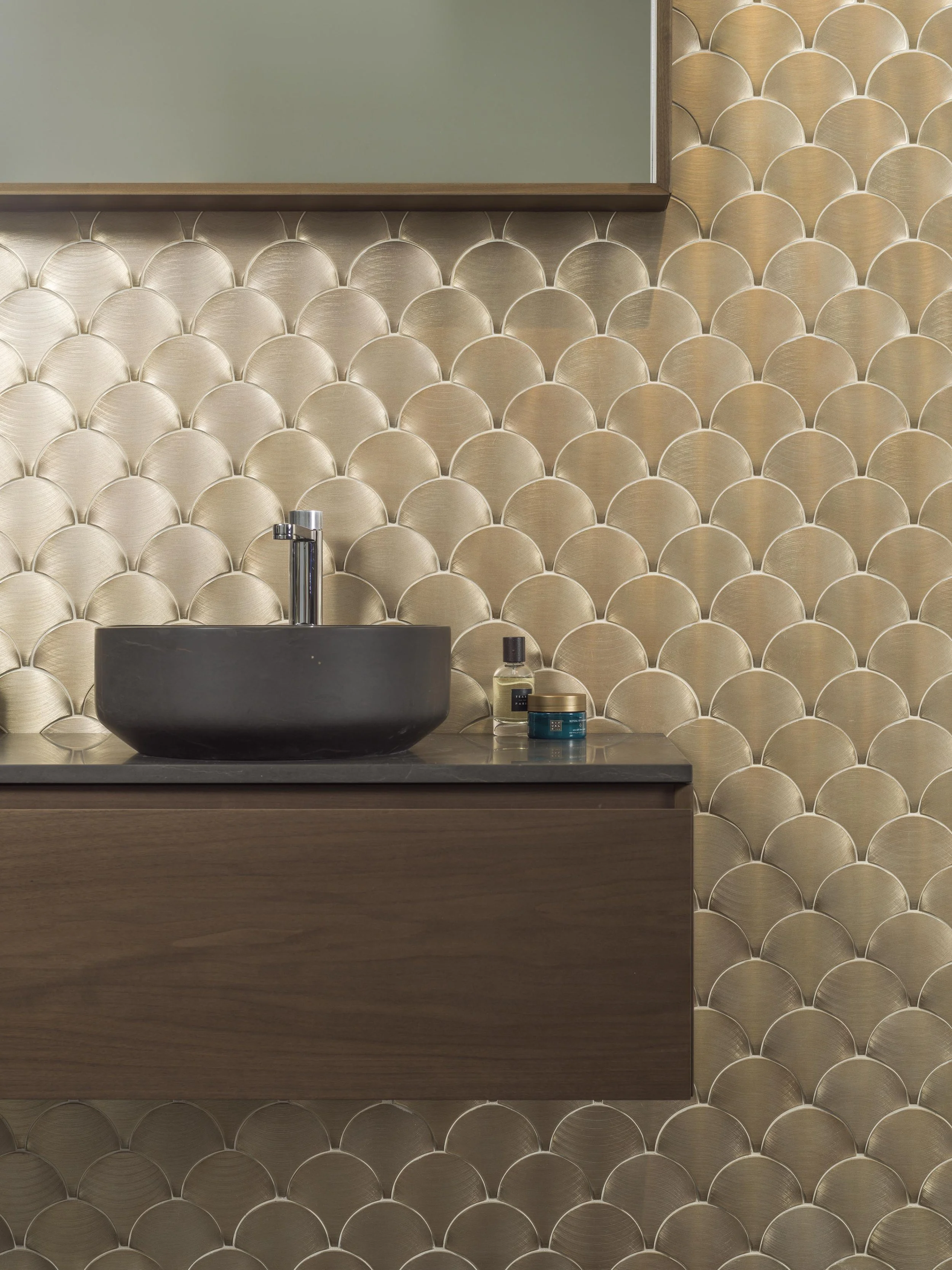 A modern bathroom sink area with a black vessel sink, a chrome faucet, and a wooden vanity. The backsplash features a gold, scalloped, tile pattern. There are bottles of toiletries on the countertop.