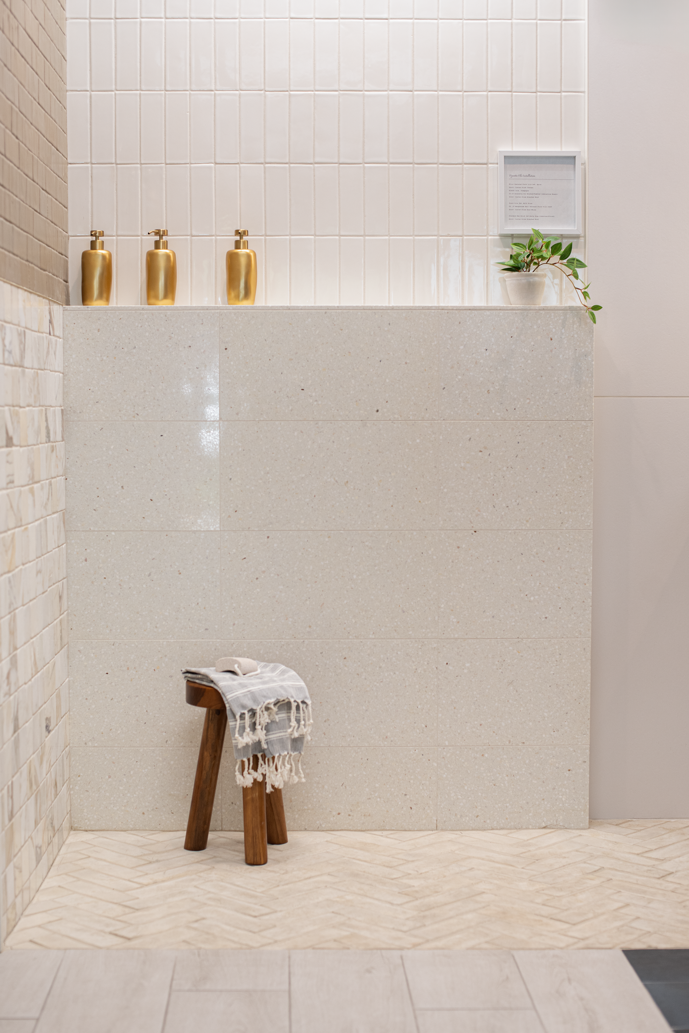 Minimalist bathroom interior with white tiled walls, a beige stone counter, three gold soap dispensers, a small framed sign, a potted green plant, a wooden stool with a striped towel, and beige herringbone-patterned floor tiles.