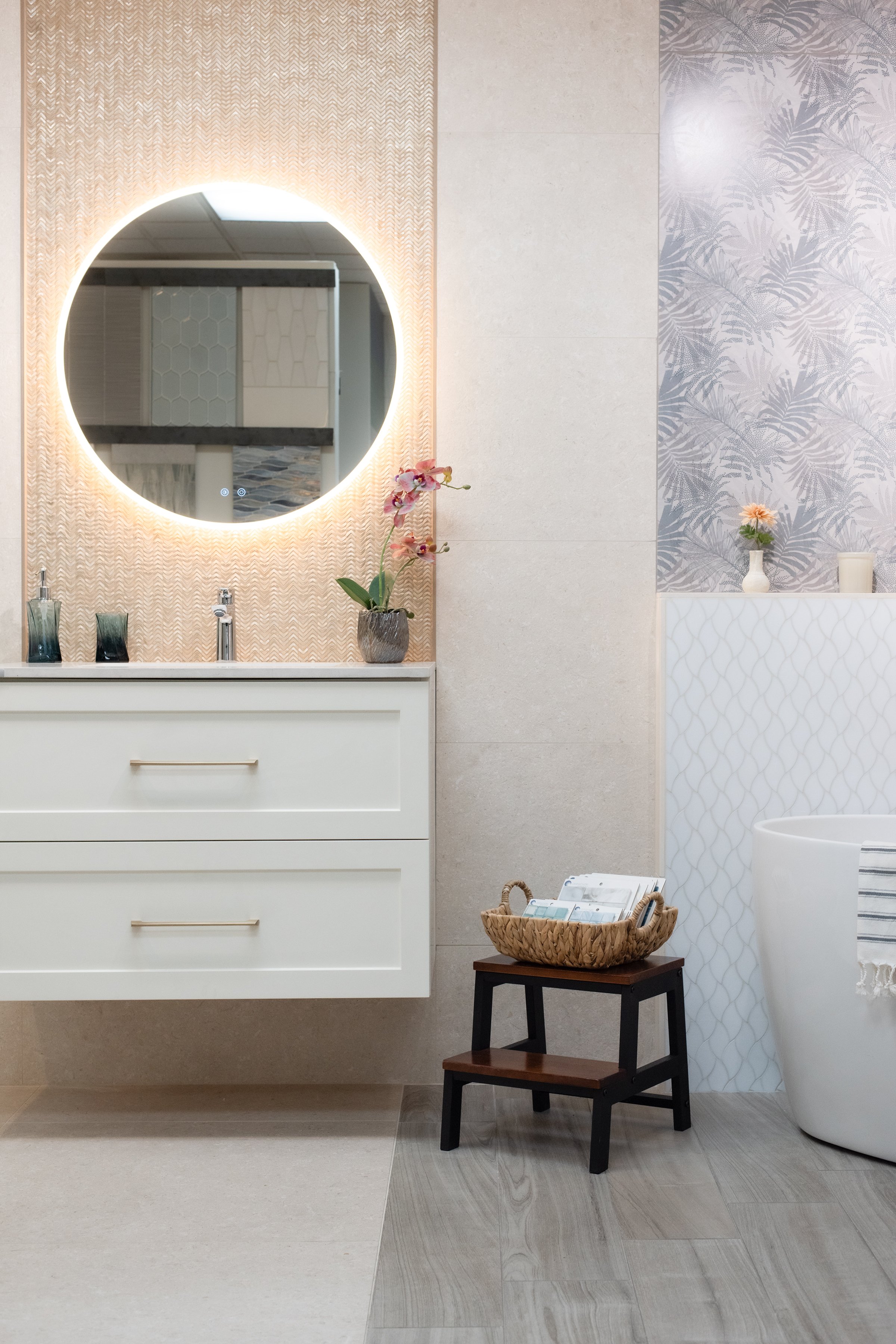 Modern bathroom with a backlit round mirror above a white vanity, a potted orchid, small decorative bottles, a small table with magazines, and a partial view of a bathtub with a towel.