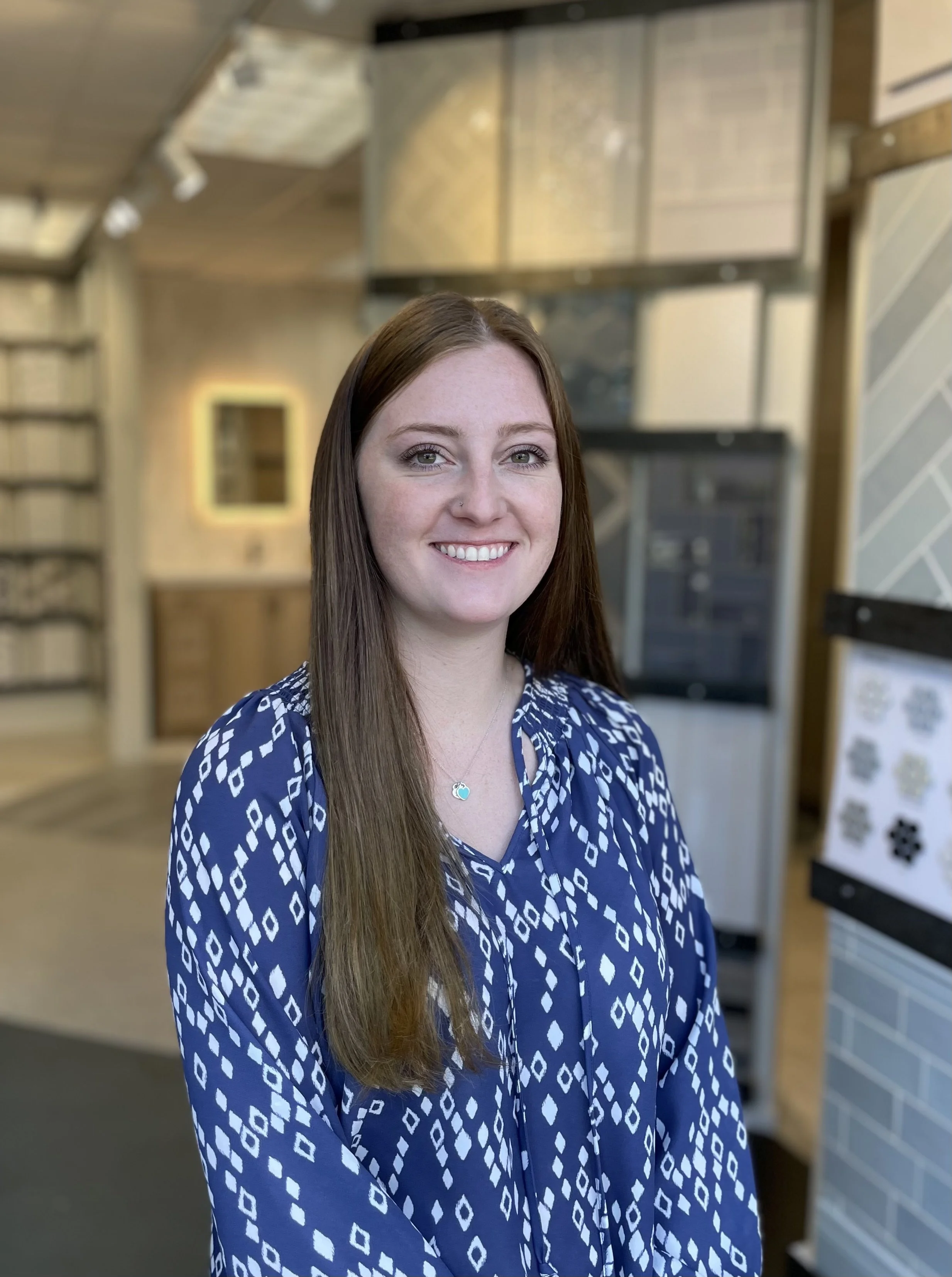 A young woman with long brown hair and a blue patterned blouse smiling, standing indoors with wooden and brick decor.