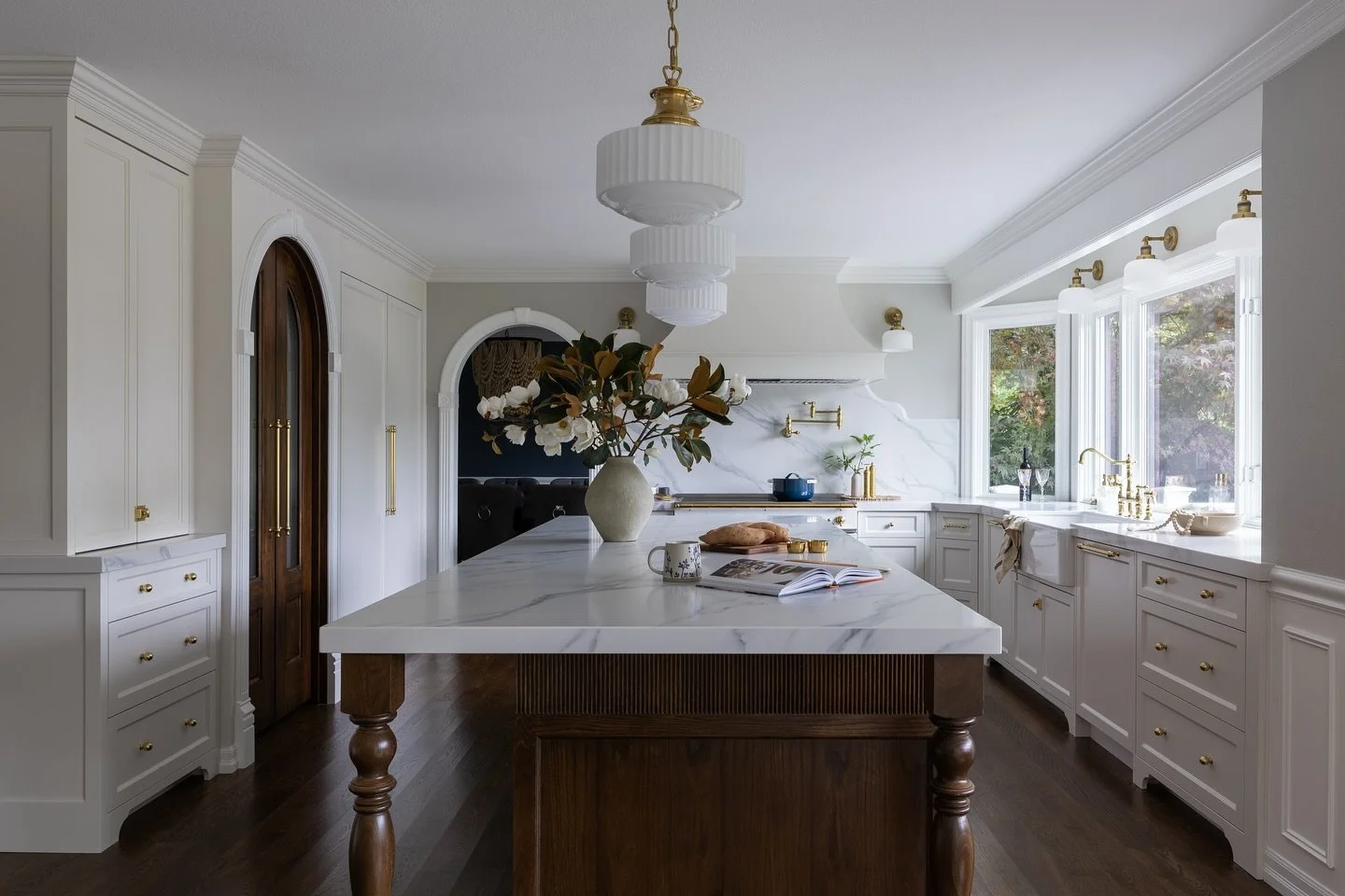 Bright modern kitchen with white cabinetry, a large central island with a marble countertop, hanging white pendant lights, and a window overlooking greenery.
