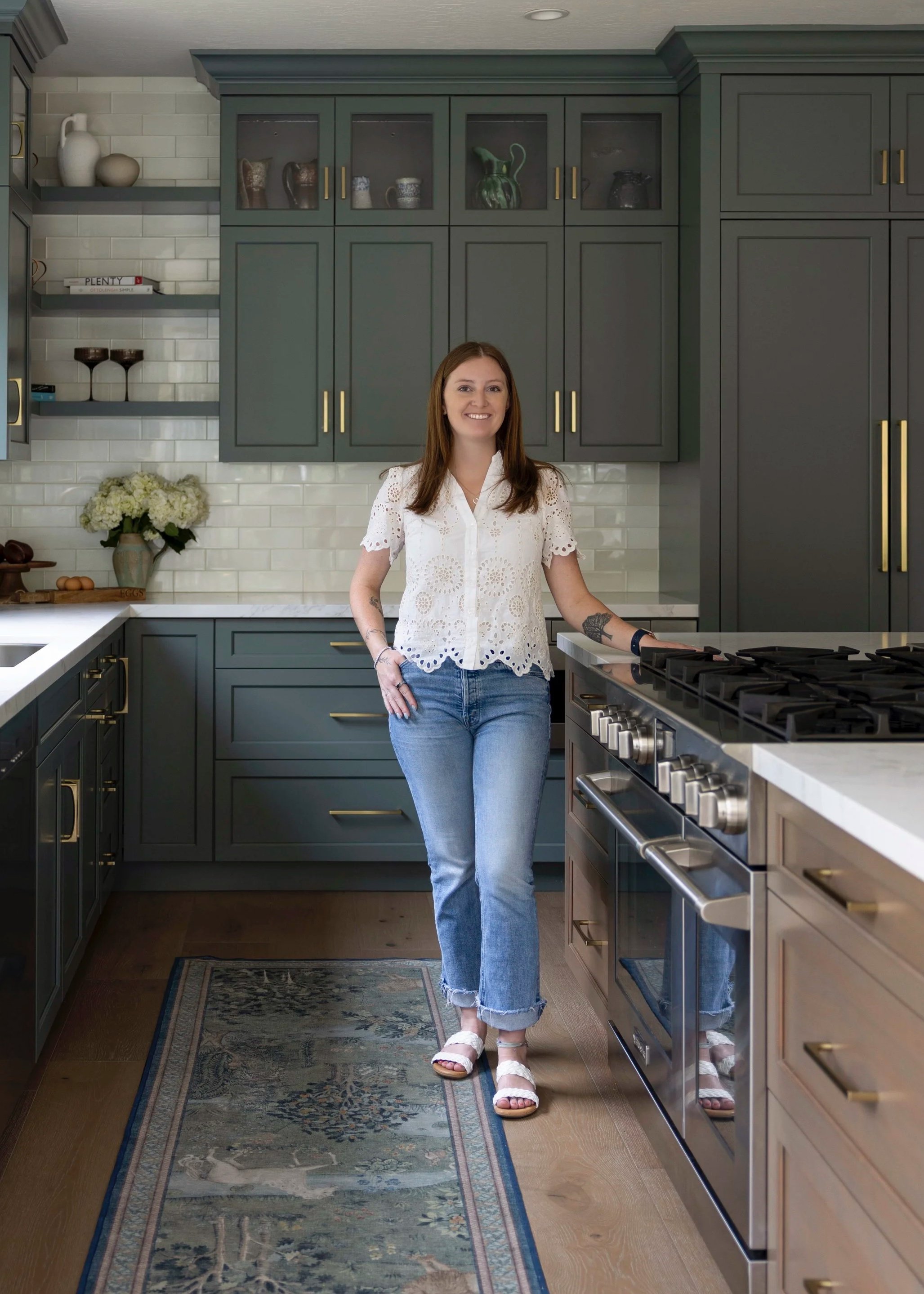 A young woman with long brown hair and a blue patterned blouse smiling, standing indoors with wooden and brick decor.