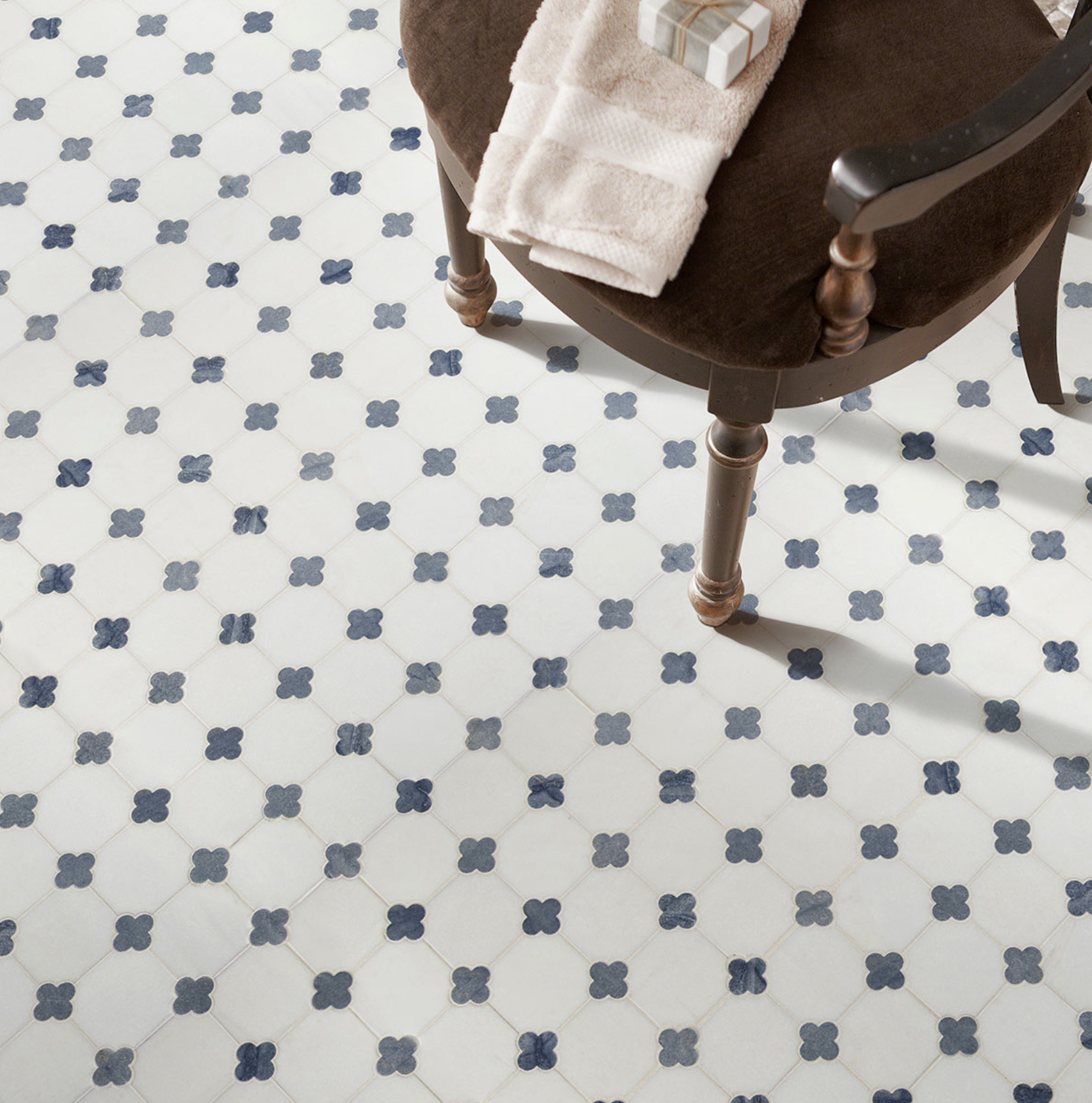 A brown upholstered chair with wooden legs, a towel, a small gift box, and a dark armrest on a patterned tile floor.