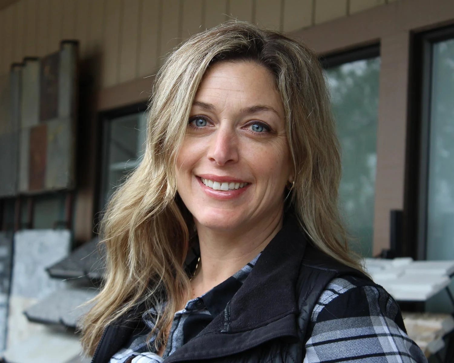 A woman with long blond hair and blue eyes smiling in front of a house with large windows and outdoor stone features.