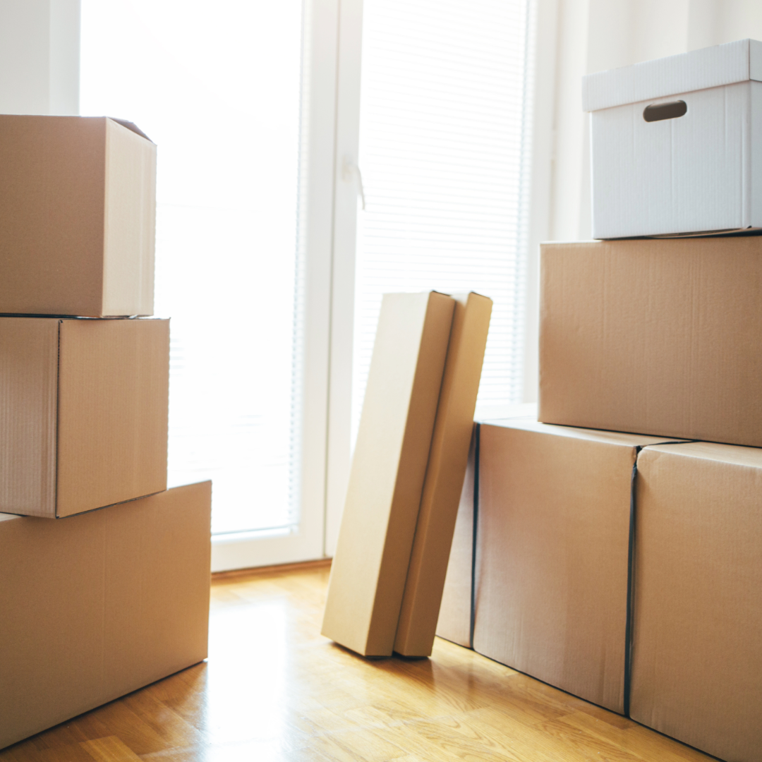 Moving boxes in an empty, sunlit room near a window and open door, suggesting a transition to what comes next.