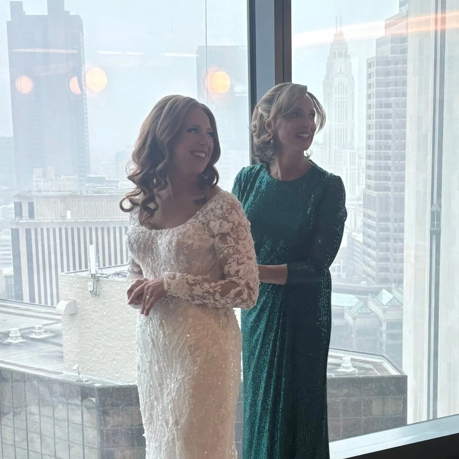 Teresa Kenney helping her daughter adjust her wedding dress near large windows overlooking the Columbus, Ohio skyline before the ceremony.