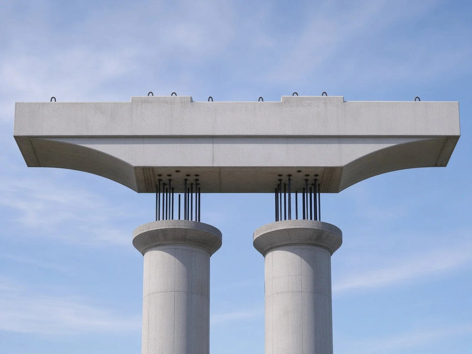 Under construction of a large concrete building with two concrete pillars and a horizontal concrete beam placed on top, set against a blue sky with clouds.