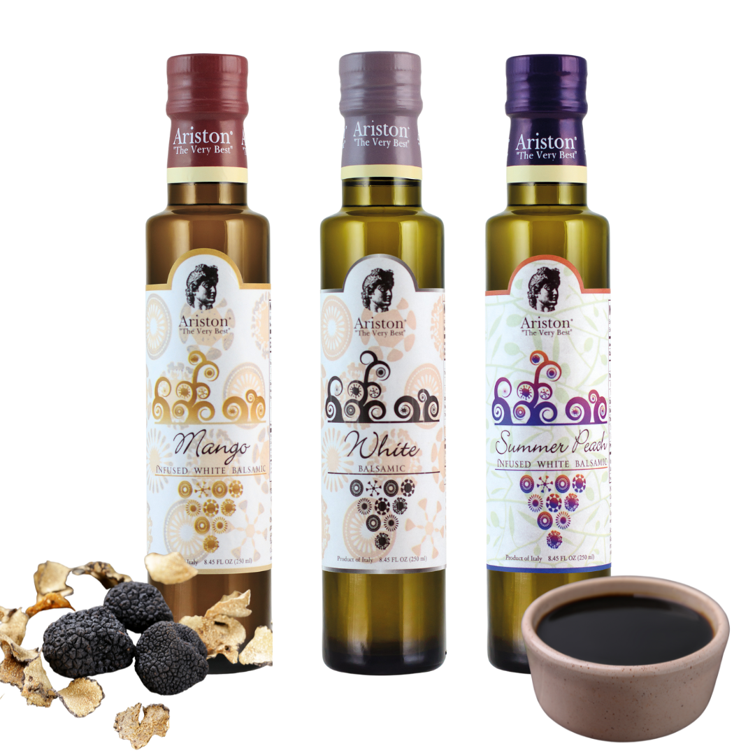 Three bottles of Ariston infused white balsamic vinegar in flavors of Mango, White, and Summer Peach, with a bowl of black vinegar and blackberries in front.