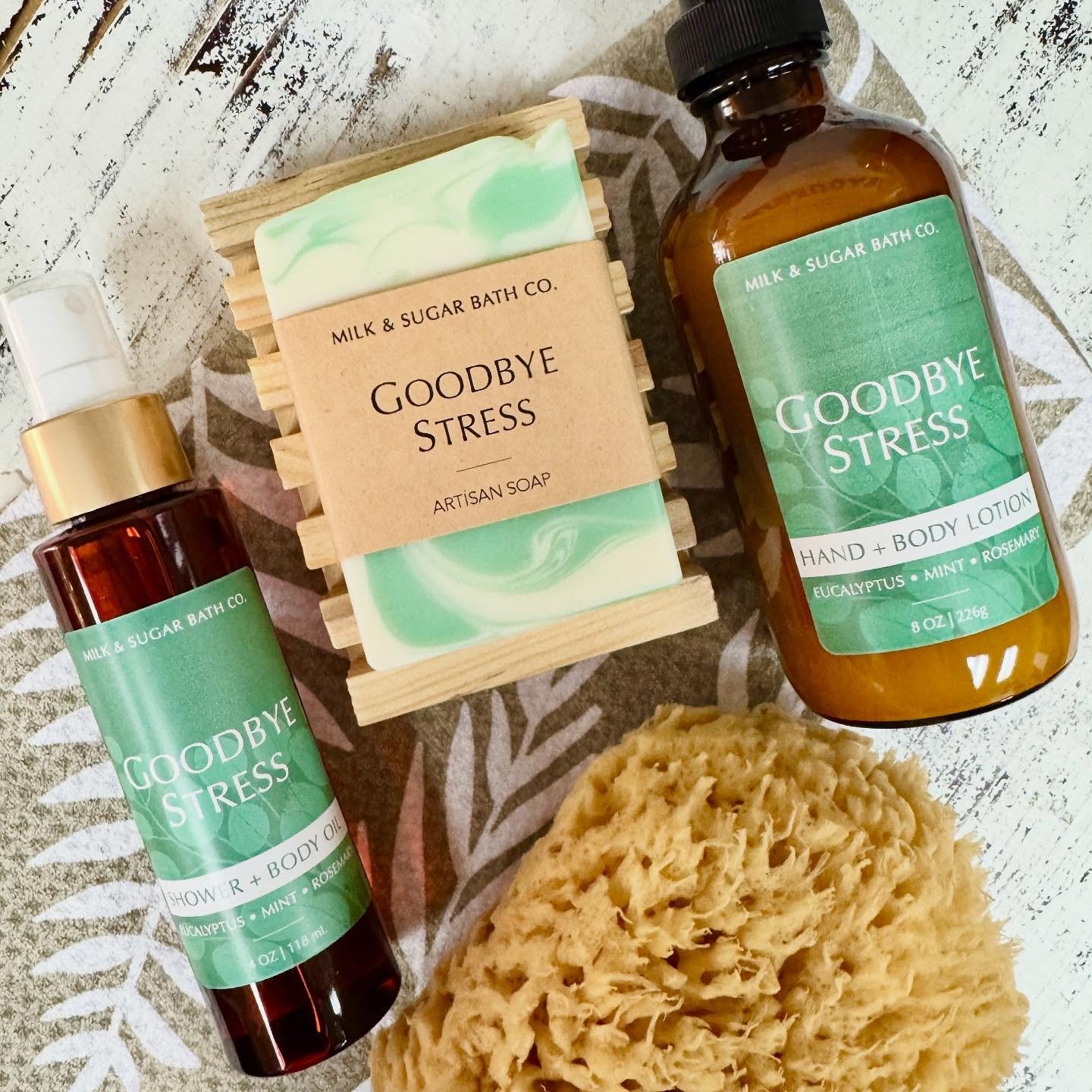 A collection of bath and body products including a bar of artisan soap labeled 'Goodbye Stress,' a bottle of hand and body lotion labeled 'Goodbye Stress,' a bottle of shower and body oil labeled 'Goodbye Stress,' a natural loofah, and a white cloth with a leaf pattern in the background.