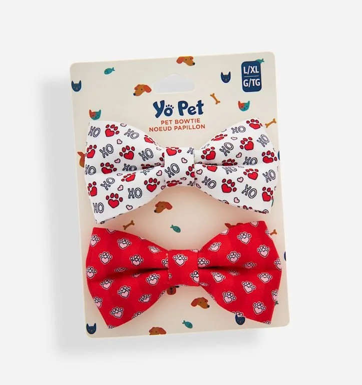 Two pet bowties, one white with red and blue paw prints and "XO" symbols, and one red with white paw prints and blue dots, attached to a cardboard backing with a dog and cat pattern.