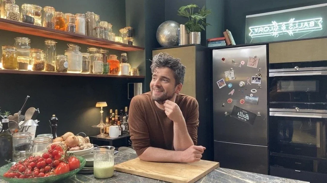 Een man die in een moderne keuken staat, omringd door keukengerei, verse tomaten, uien, een glas sap, en een koelkast met magneten en foto's, terwijl hij glimlacht.