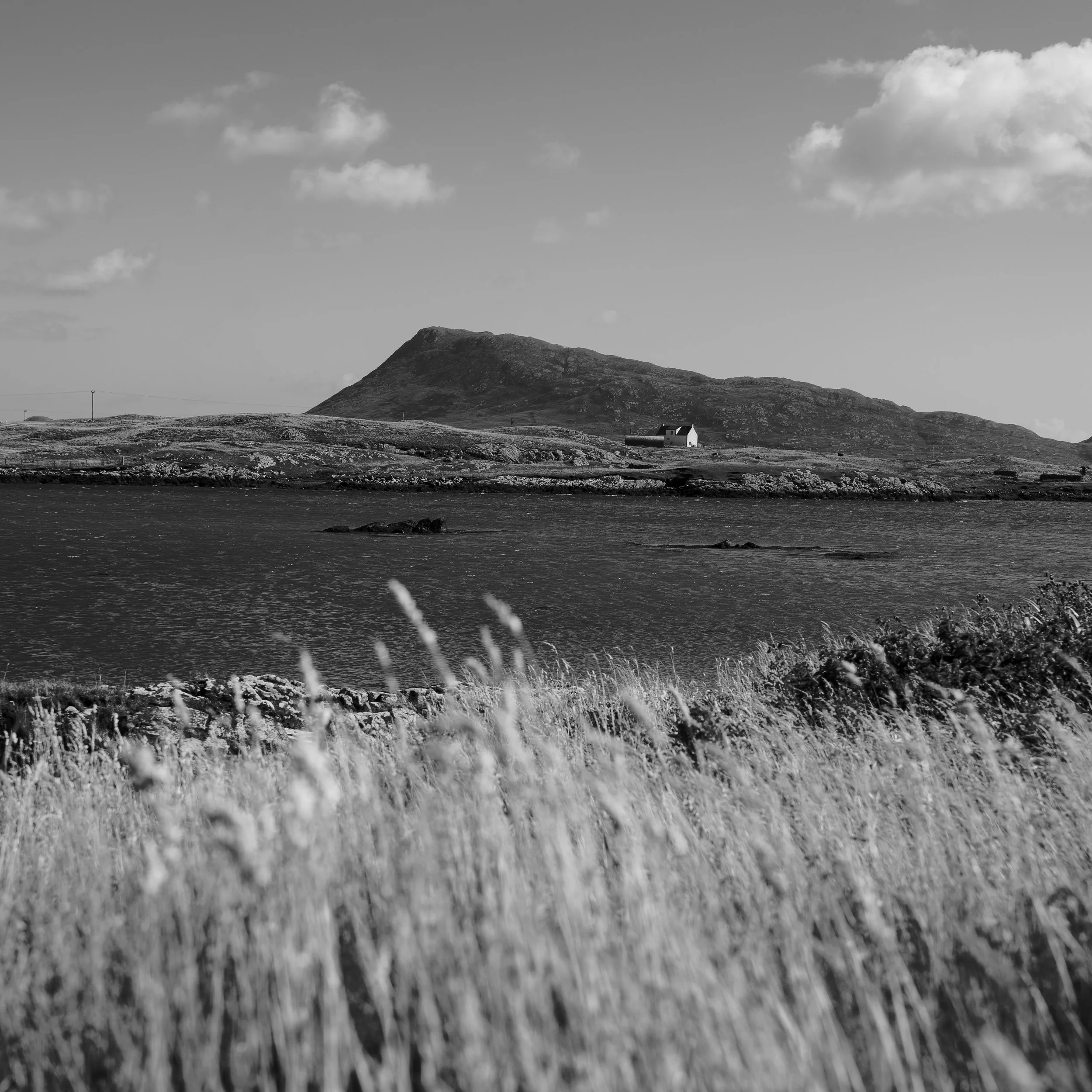 A black and white landscape photograph of a mountain with a small white house at its base, a body of water in the foreground, and a sky with scattered clouds.
