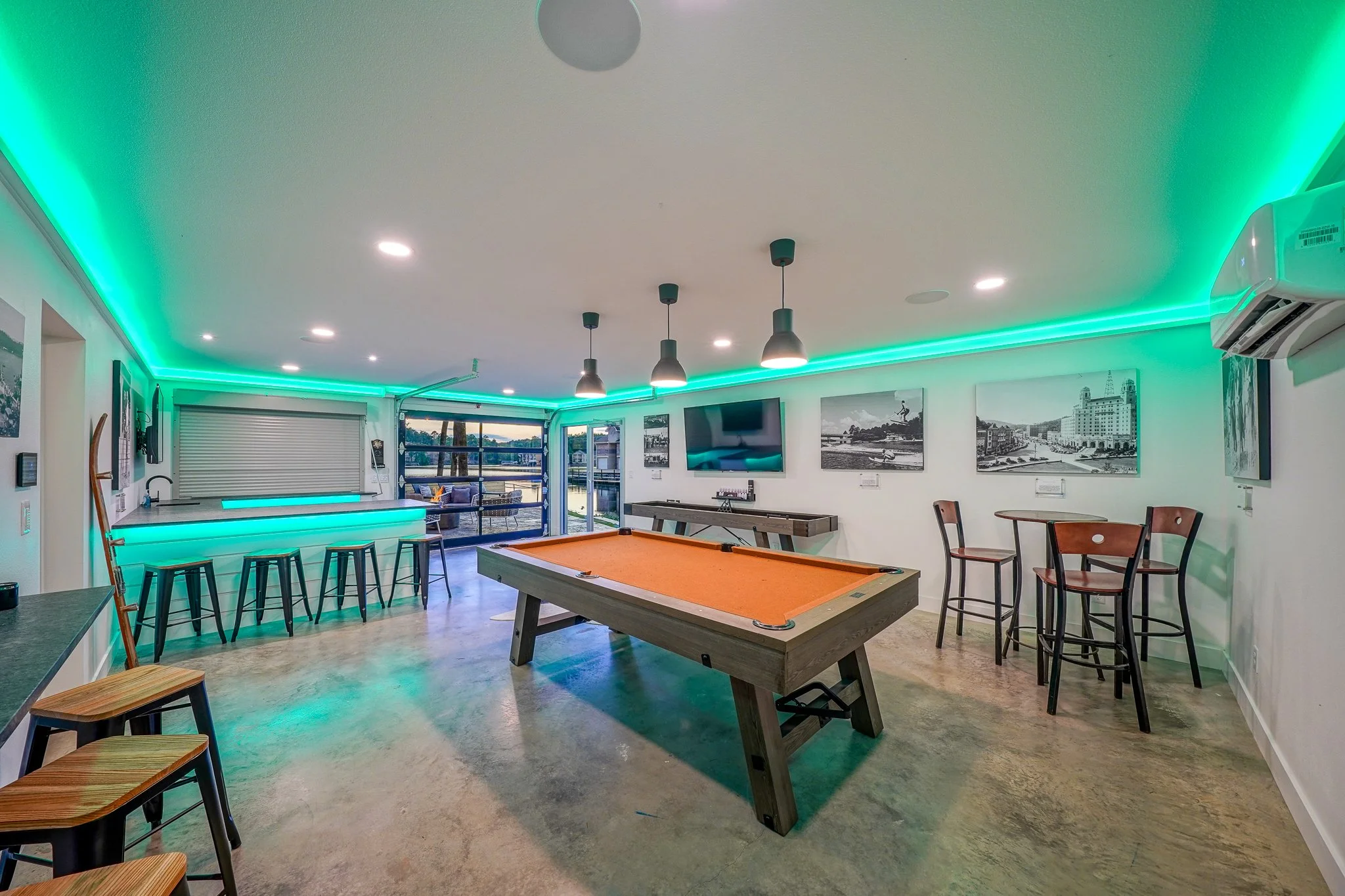  A spacious game room featuring a pool table, a bar, and bar and table seating. The room is illuminated with green LED strip lighting along the ceiling.