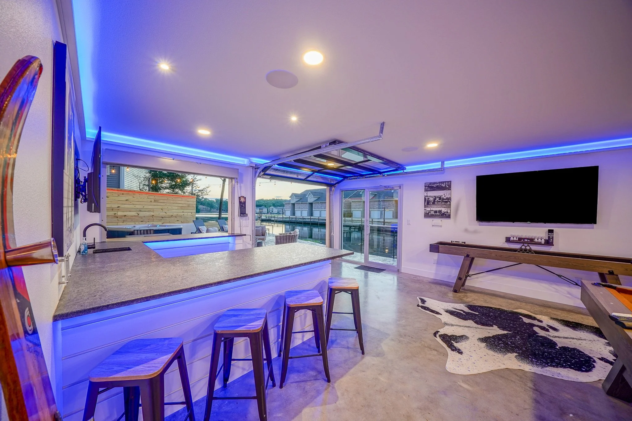 A modern entertainment room with a large countertop bar area and a large flat-screen TV. The room is illuminated with blue LED strip lighting along the ceiling.