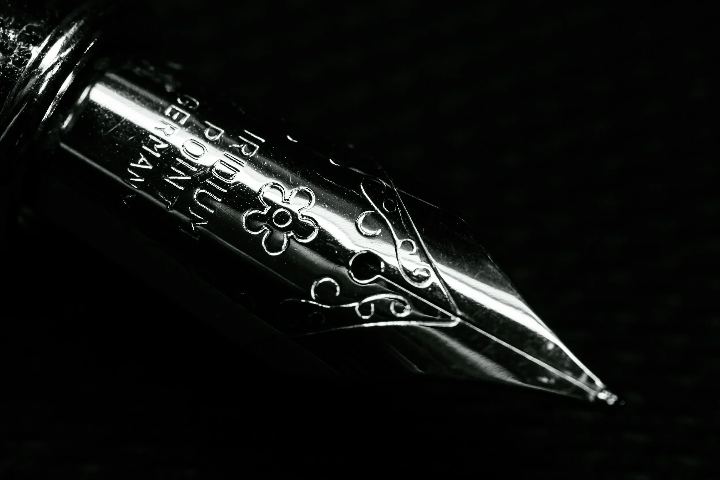 Close-up of a shiny, metallic fountain pen nib with engraved decorative patterns, set against a dark background.