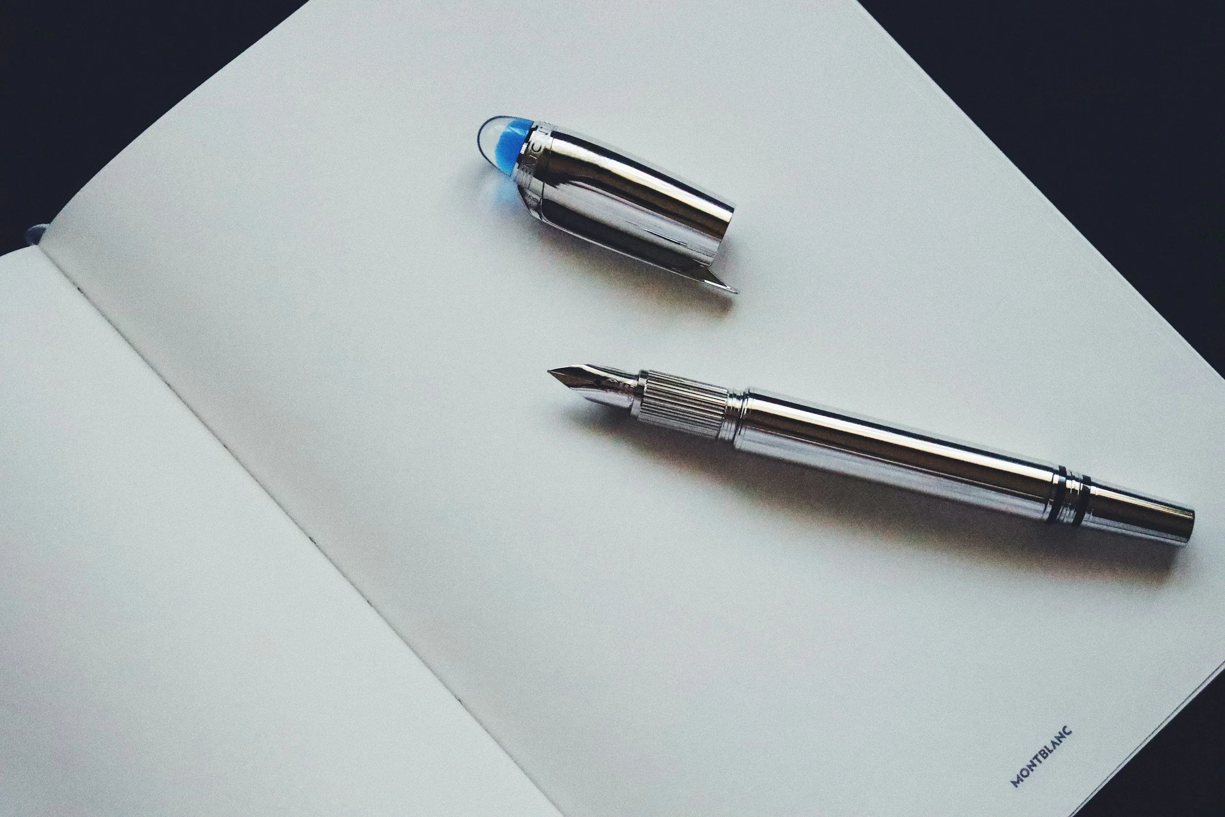 A silver fountain pen and matching cap resting on a blank sheet of white paper in an open notebook, with the brand name Montblanc printed on the bottom of the page.
