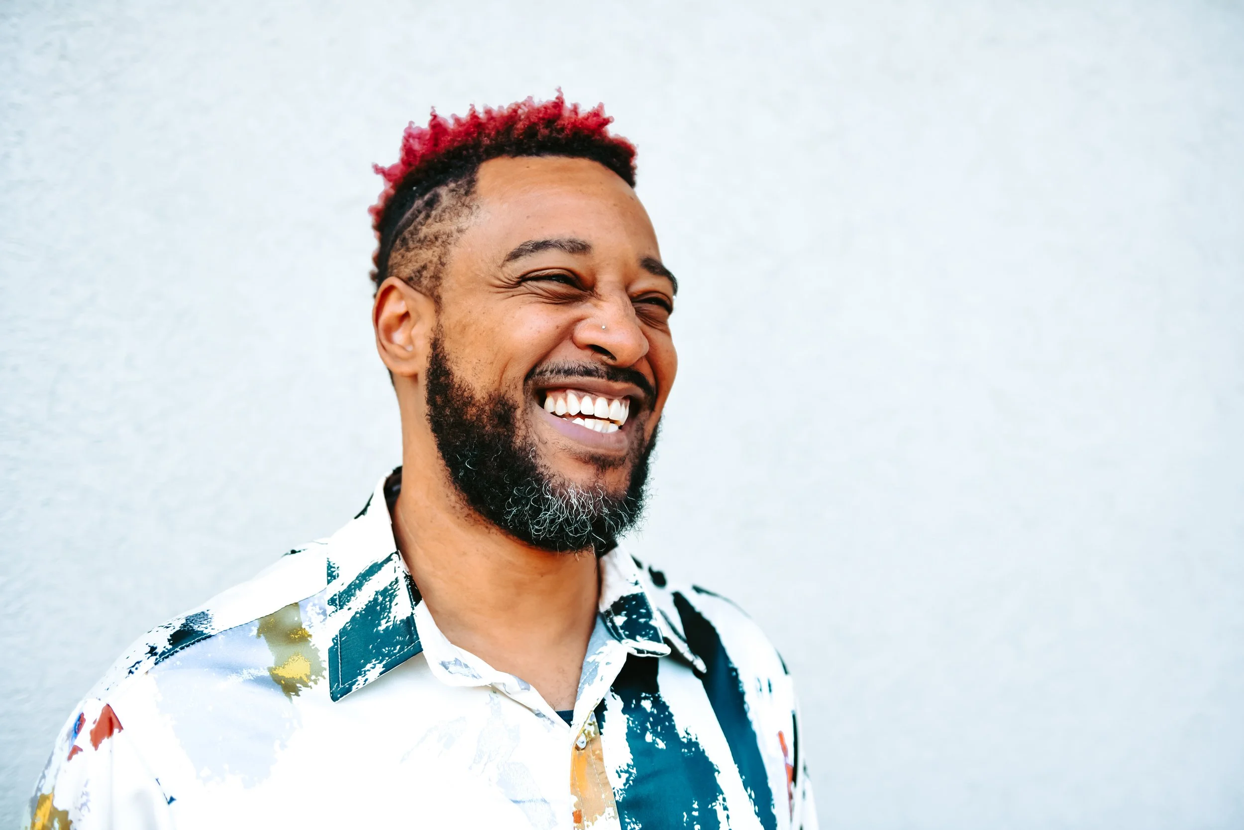 A smiling man with a beard and short hair dyed red on top, wearing a white shirt with a colorful pattern, standing against a plain light-colored background.
