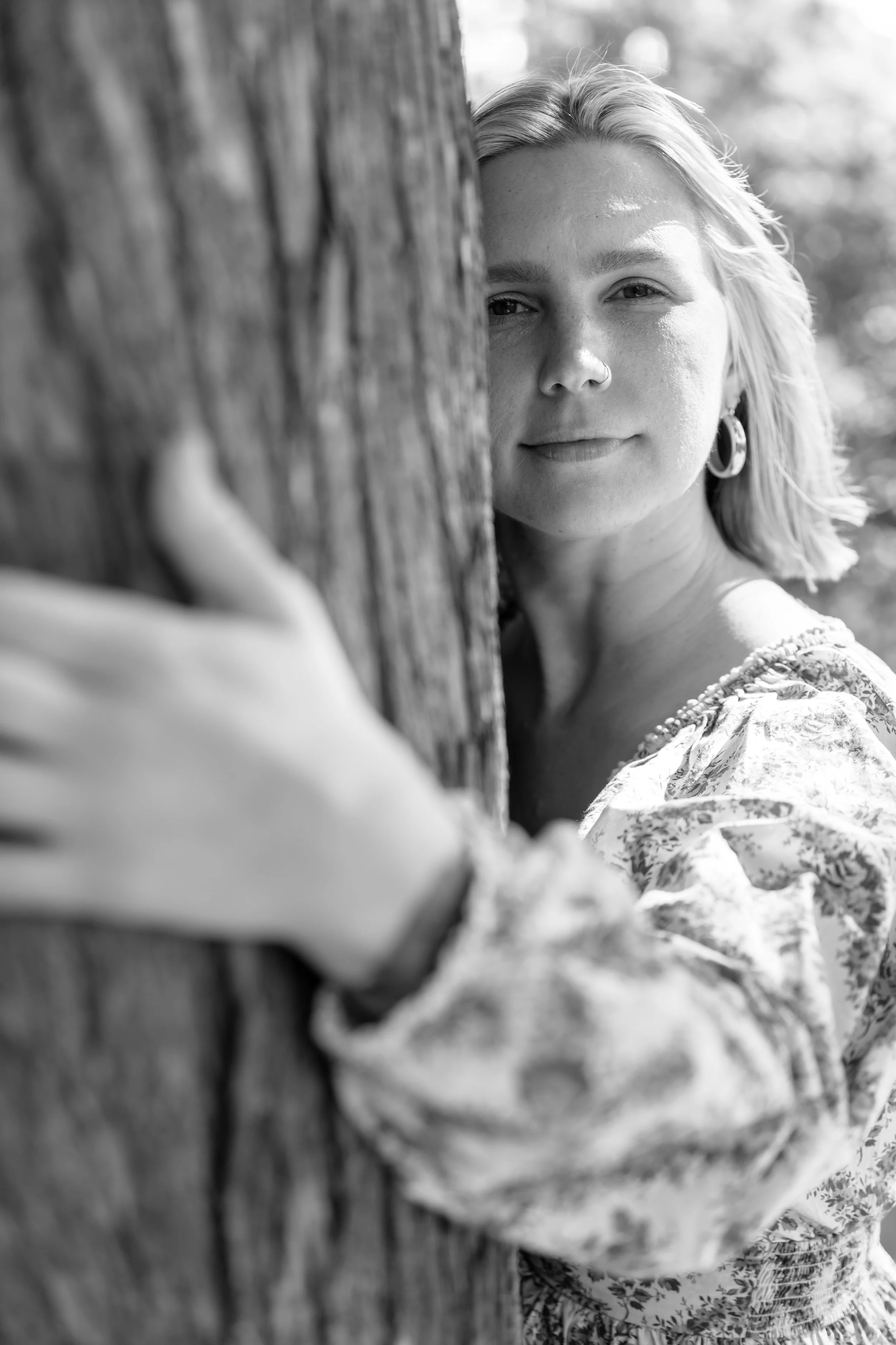A woman with shoulder-length blonde hair, wearing earrings and a floral-patterned blouse, is partially hidden behind a tree trunk, gazing towards the camera with a calm expression.