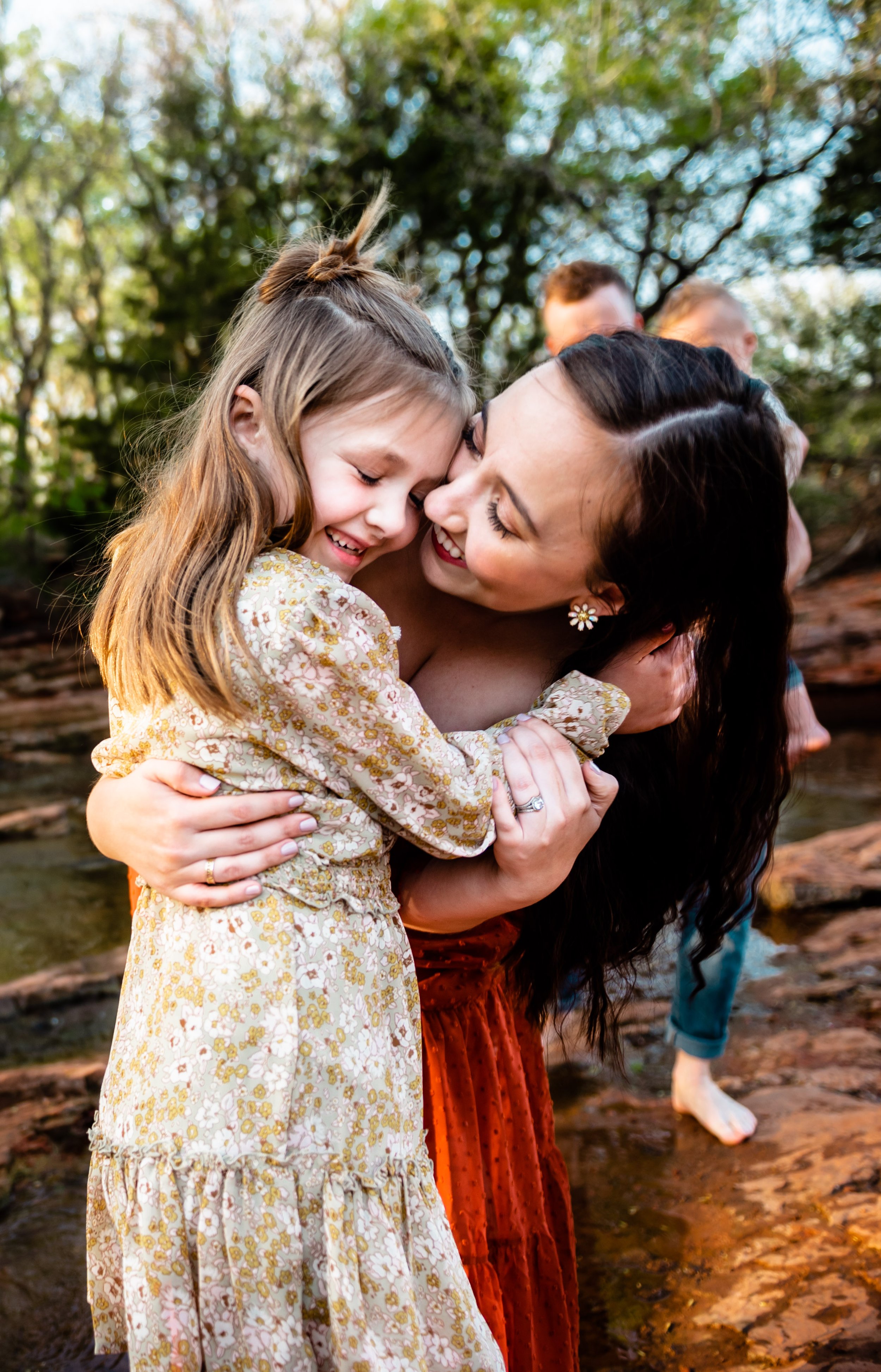 A woman with dark hair and earrings happily hugging a young girl with light brown hair, both smiling and touching foreheads outdoors with trees in the background.