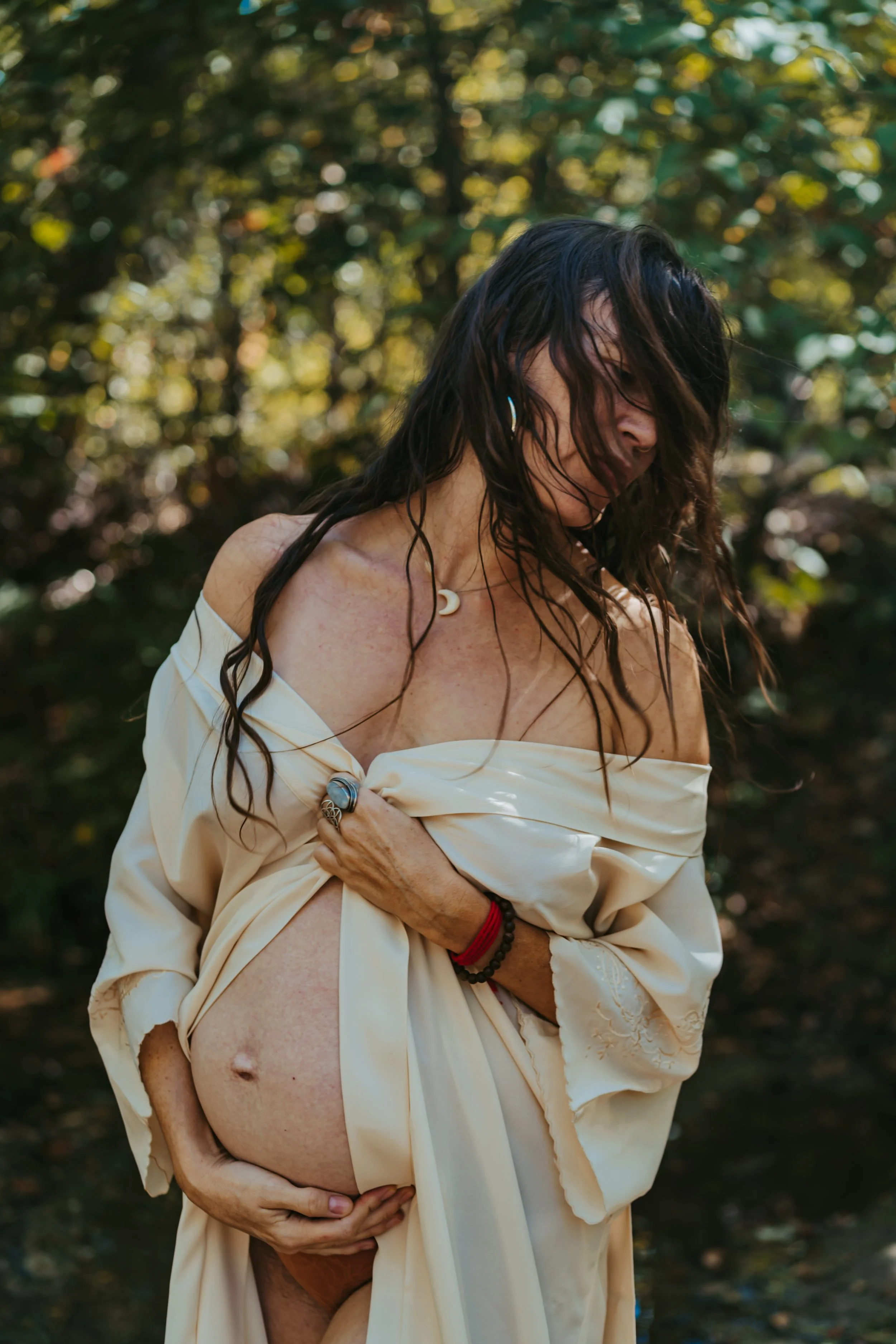 A pregnant woman with dark, wavy hair stands outdoors in a forest. She wears a cream-colored satin robe off her shoulders, exposing her belly, and has various jewelry including rings, bracelets, and a necklace with a crescent moon pendant.