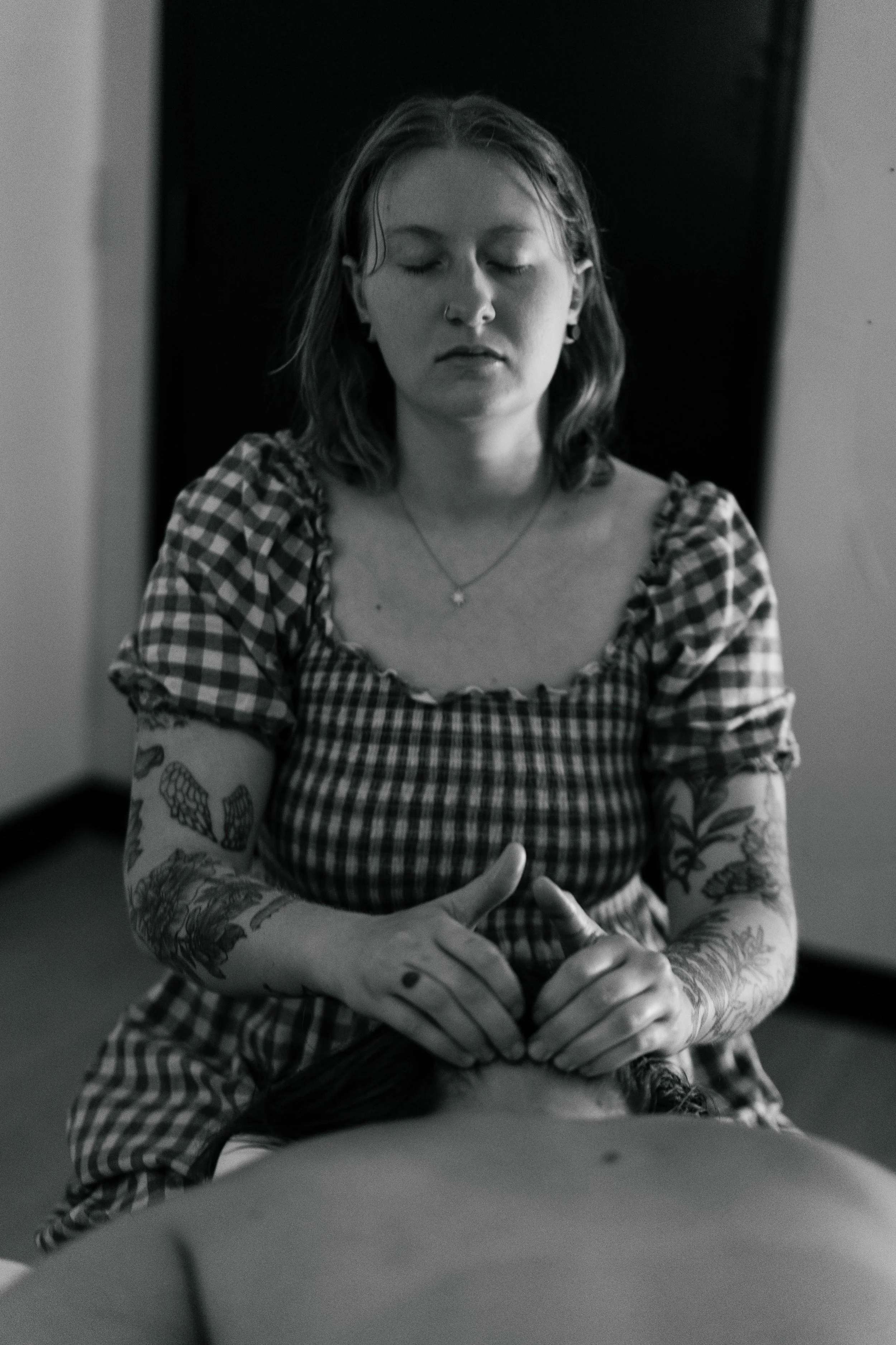 Black and white photograph of a woman with tattoos on her arms, wearing a checkered dress, sitting with her eyes closed and hands clasped on a table.