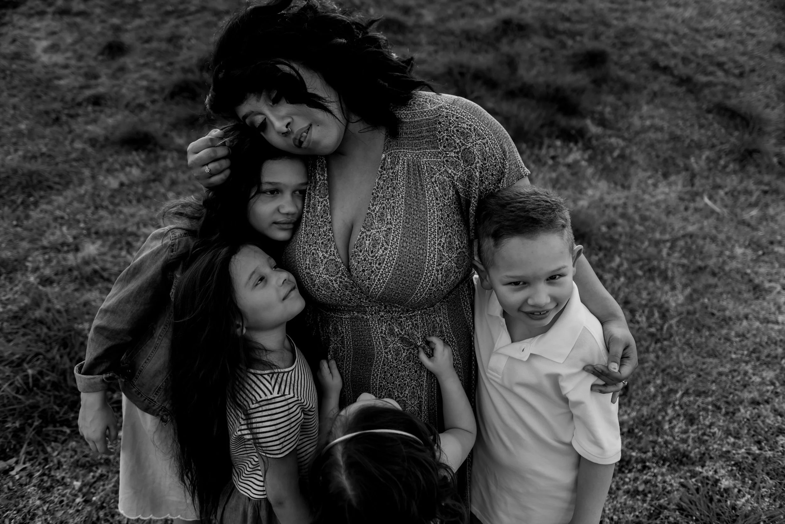 A woman with dark hair and a patterned dress hugging four children on a grassy field, black and white photo.