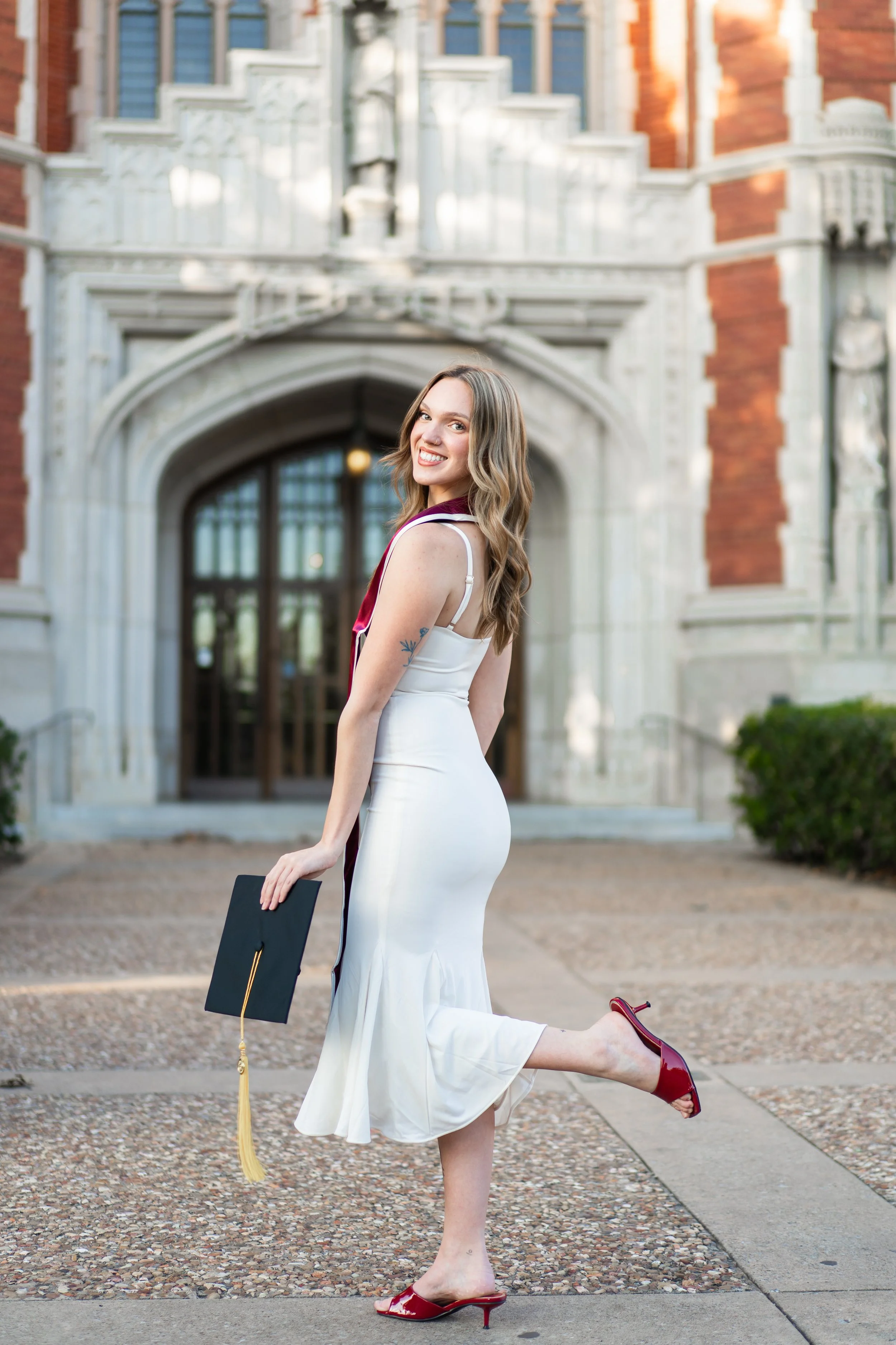 A young woman in a white dress and red high heels holding a graduation cap, standing in front of a historic building, celebrating graduation.