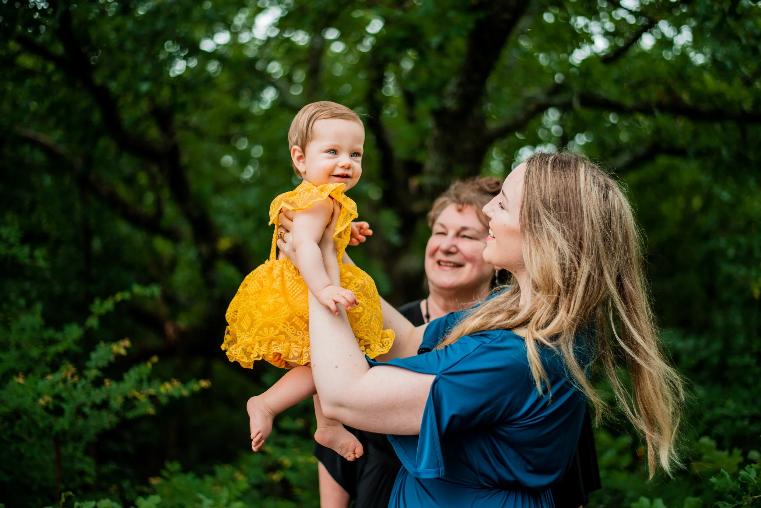 A woman holding a smiling toddler girl in a yellow dress outside with a woman in the background smiling at them.