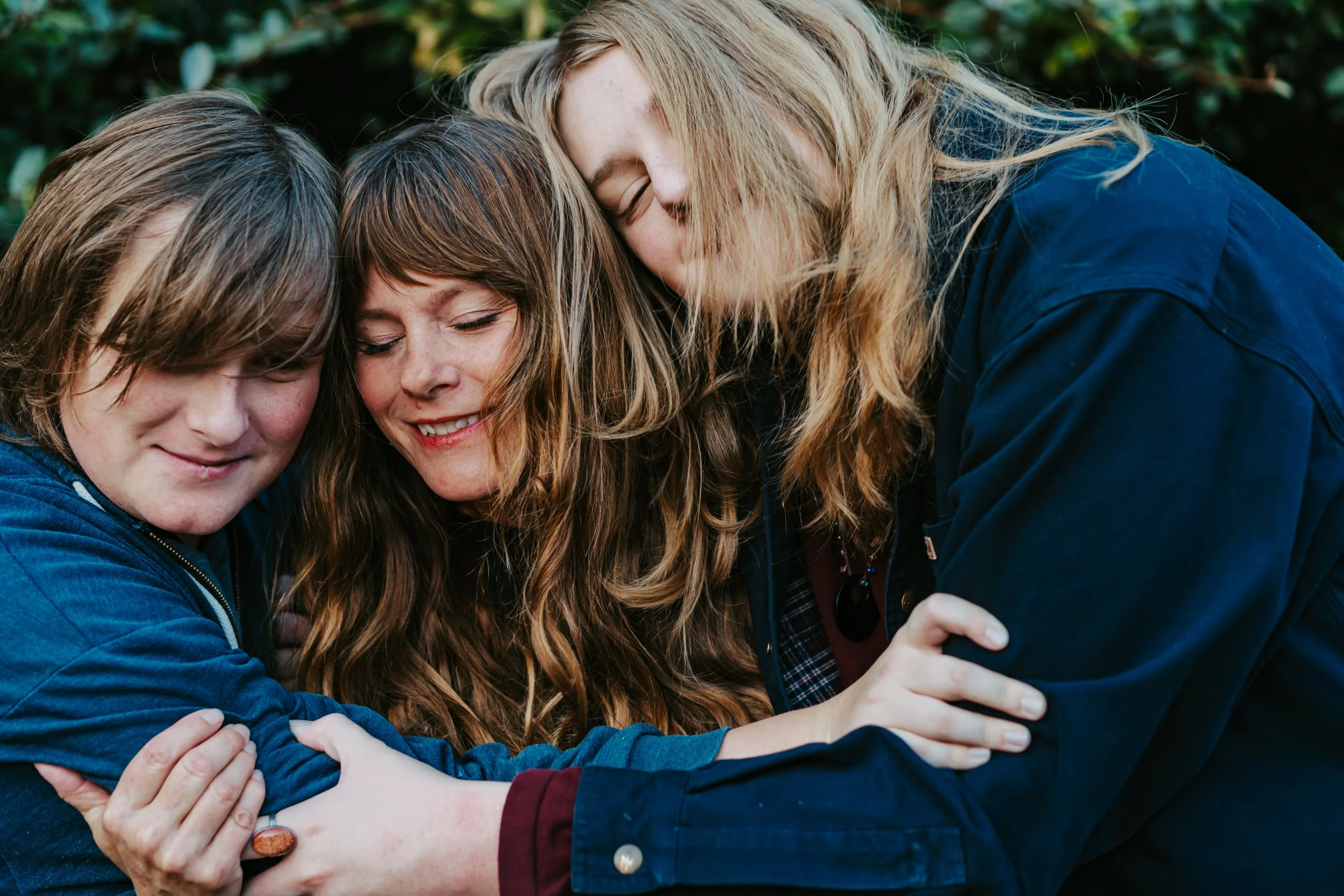 Three people sharing a group hug outdoors, smiling with eyes closed, surrounded by greenery.