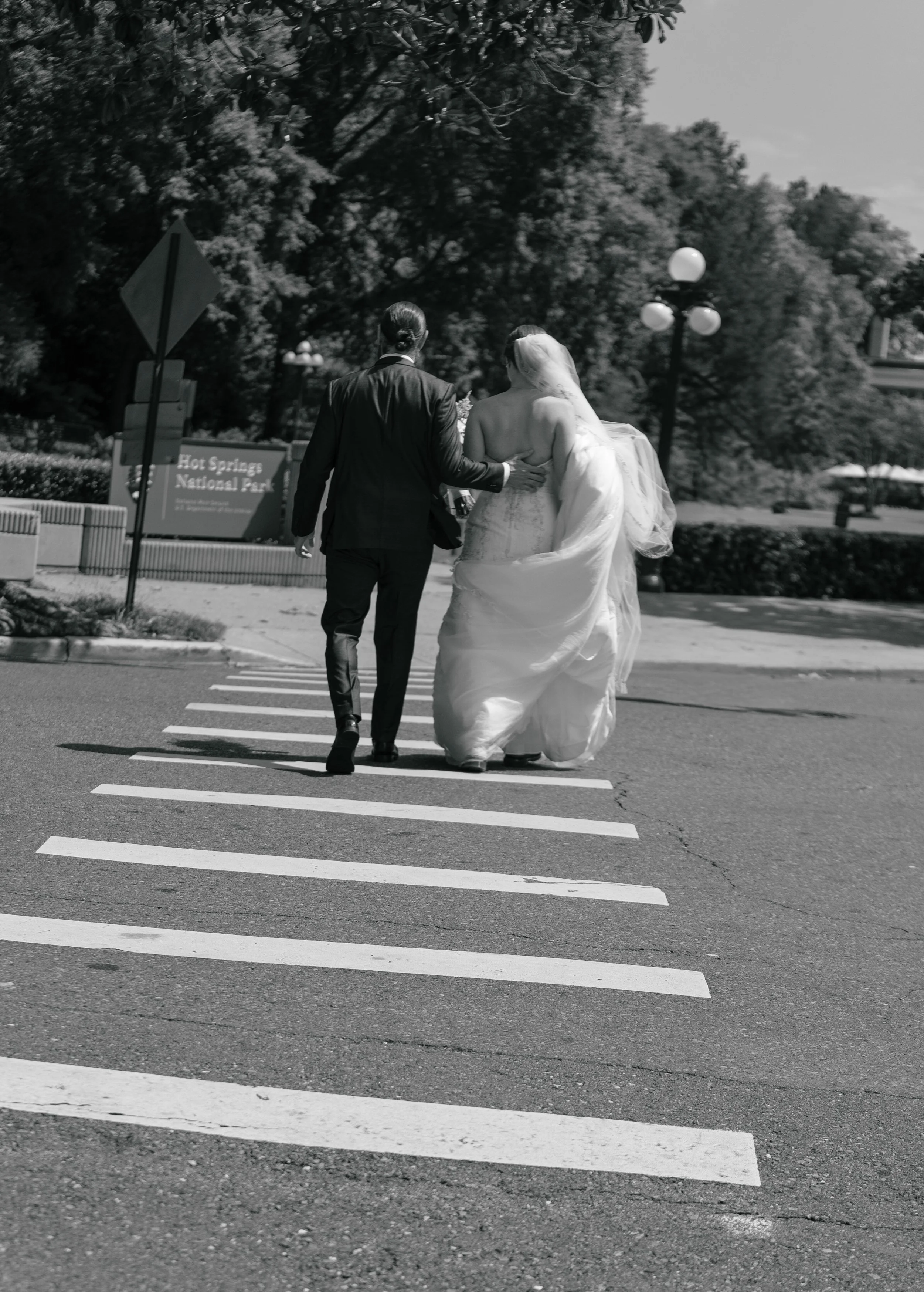 A bride and groom walking on a crosswalk in a park, with the groom in a suit and the bride in a wedding gown and veil, seen from behind in black and white.