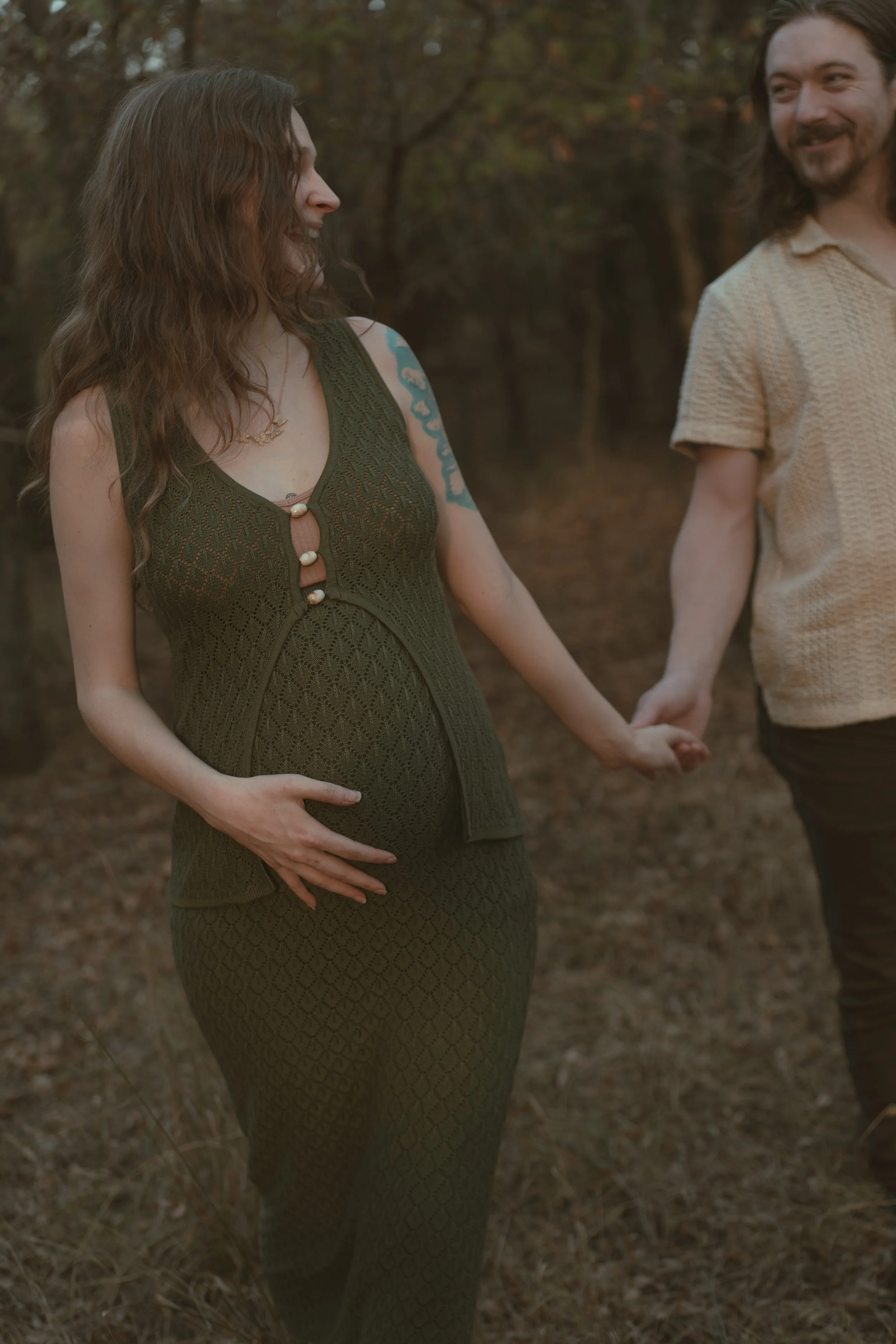 A pregnant woman wearing a green dress holding her belly, walking with a man holding her hand, outdoors on a fall day.