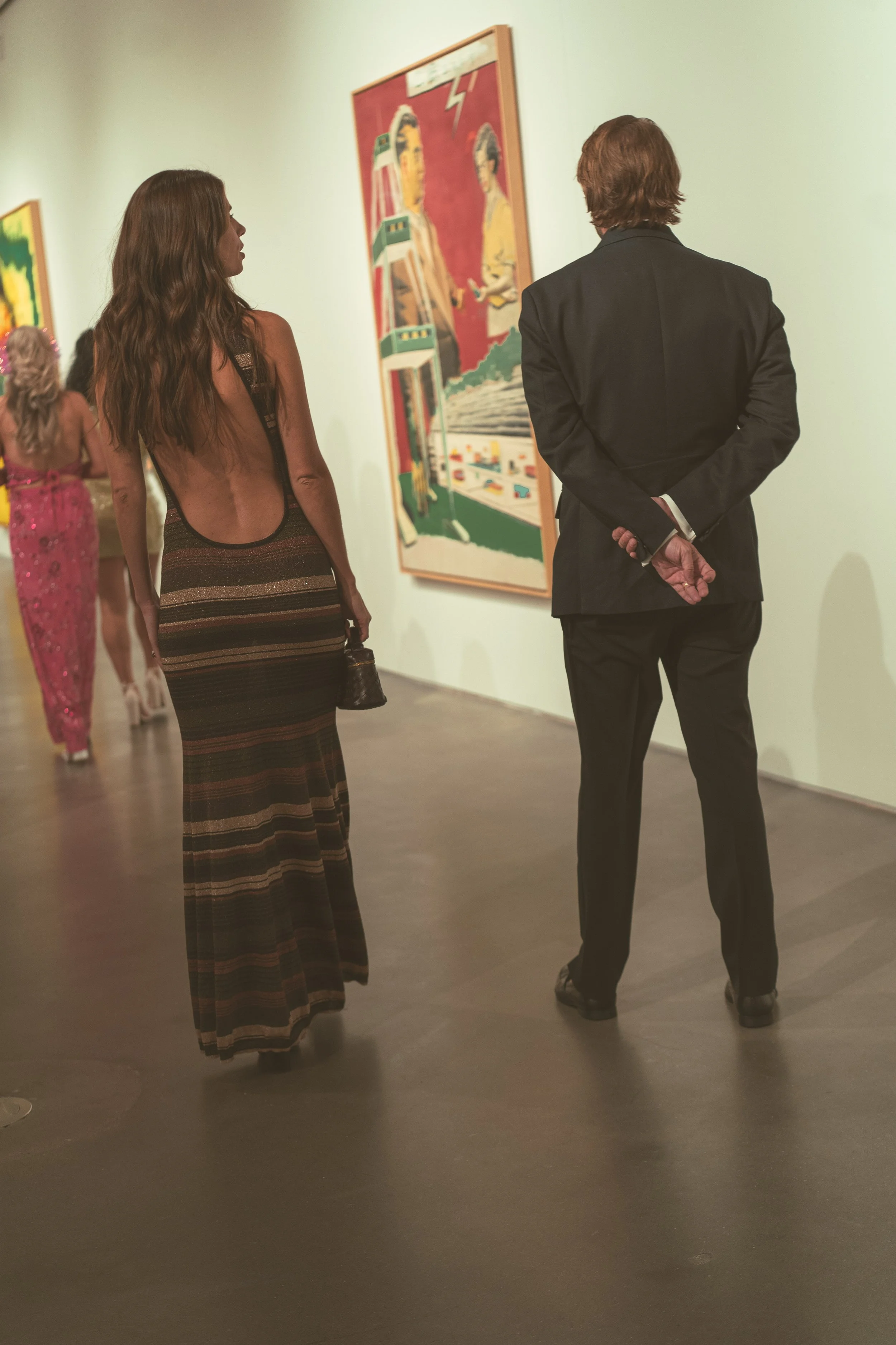 A woman with long wavy brown hair wearing a backless, striped evening gown is looking at a man in a black suit with no tie, who has his hands clasped behind his back. They are in an art gallery, viewing a colorful painting of two women in traditional attire.