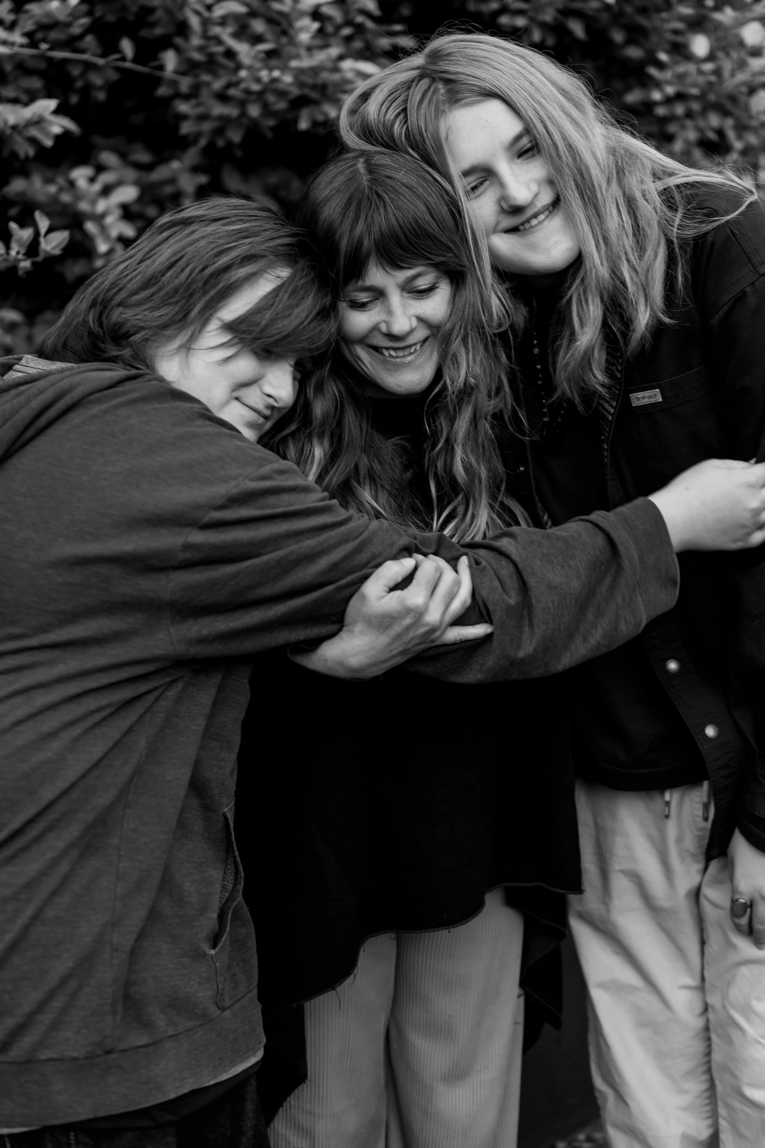 Three people hugging each other and smiling outdoors, surrounded by trees or bushes.