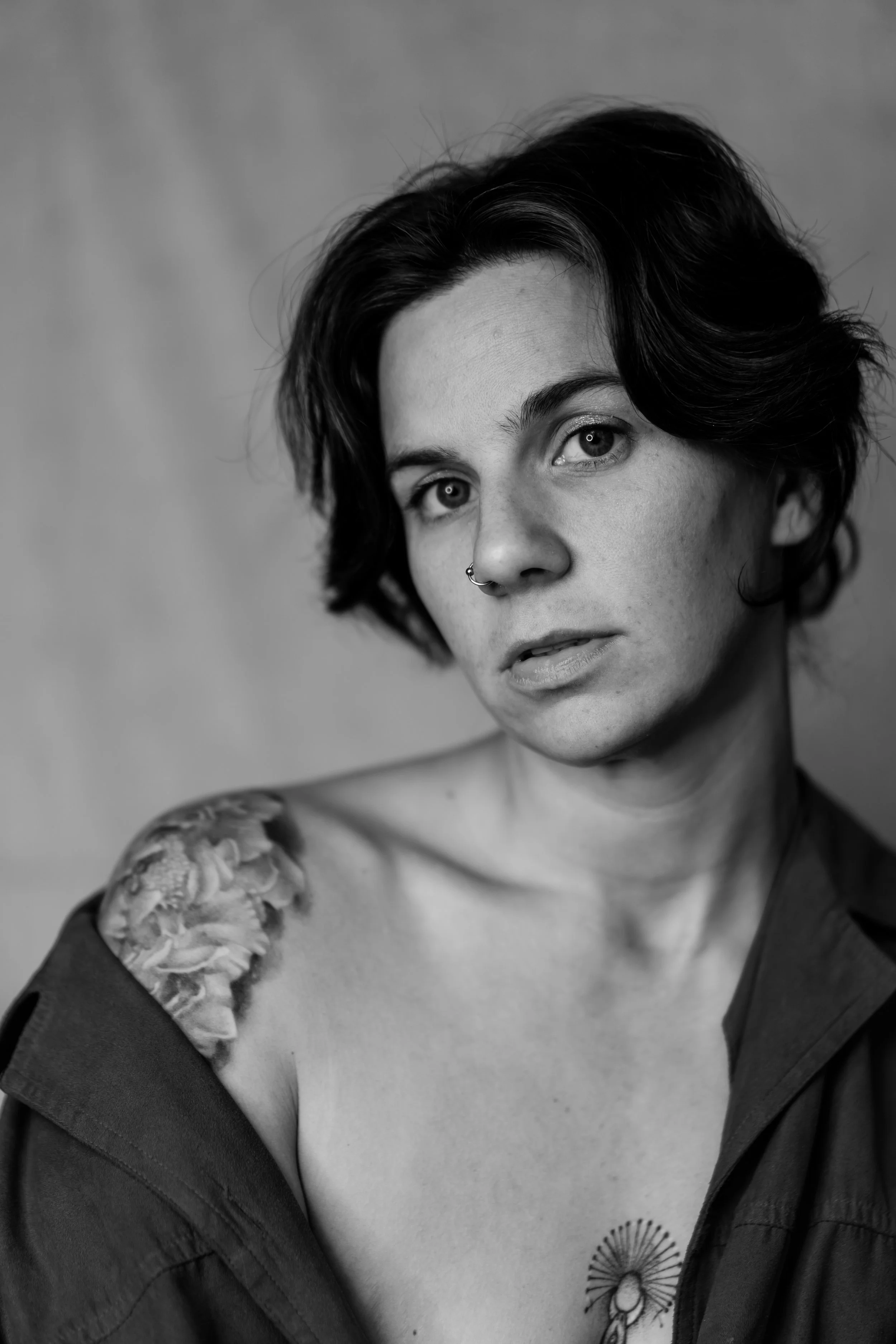 Black and white portrait of a woman with short, wavy hair, a nose ring, and a tattoo on her shoulder, wearing an open shirt.