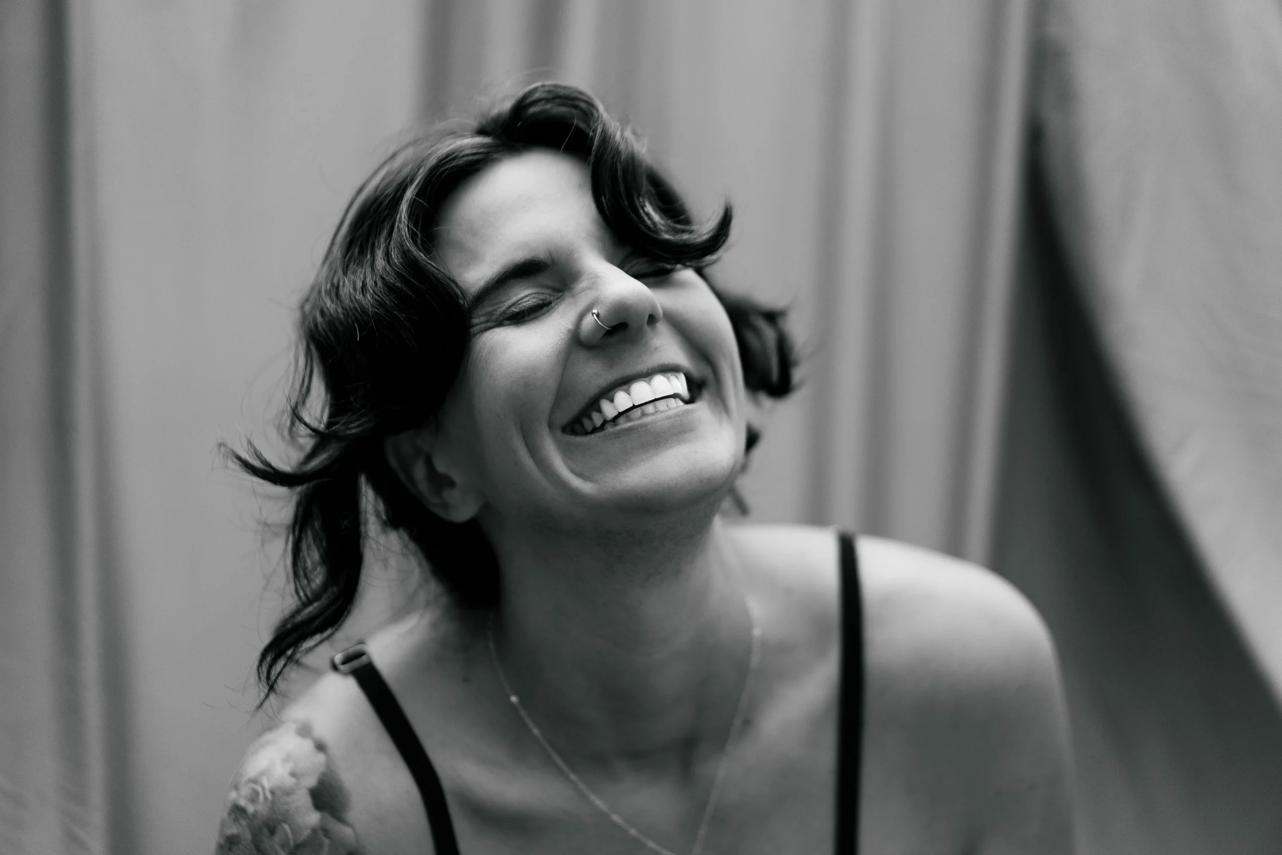 Black and white photo of a woman smiling broadly with eyes closed, short wavy hair, wearing a tank top, and a nose ring.
