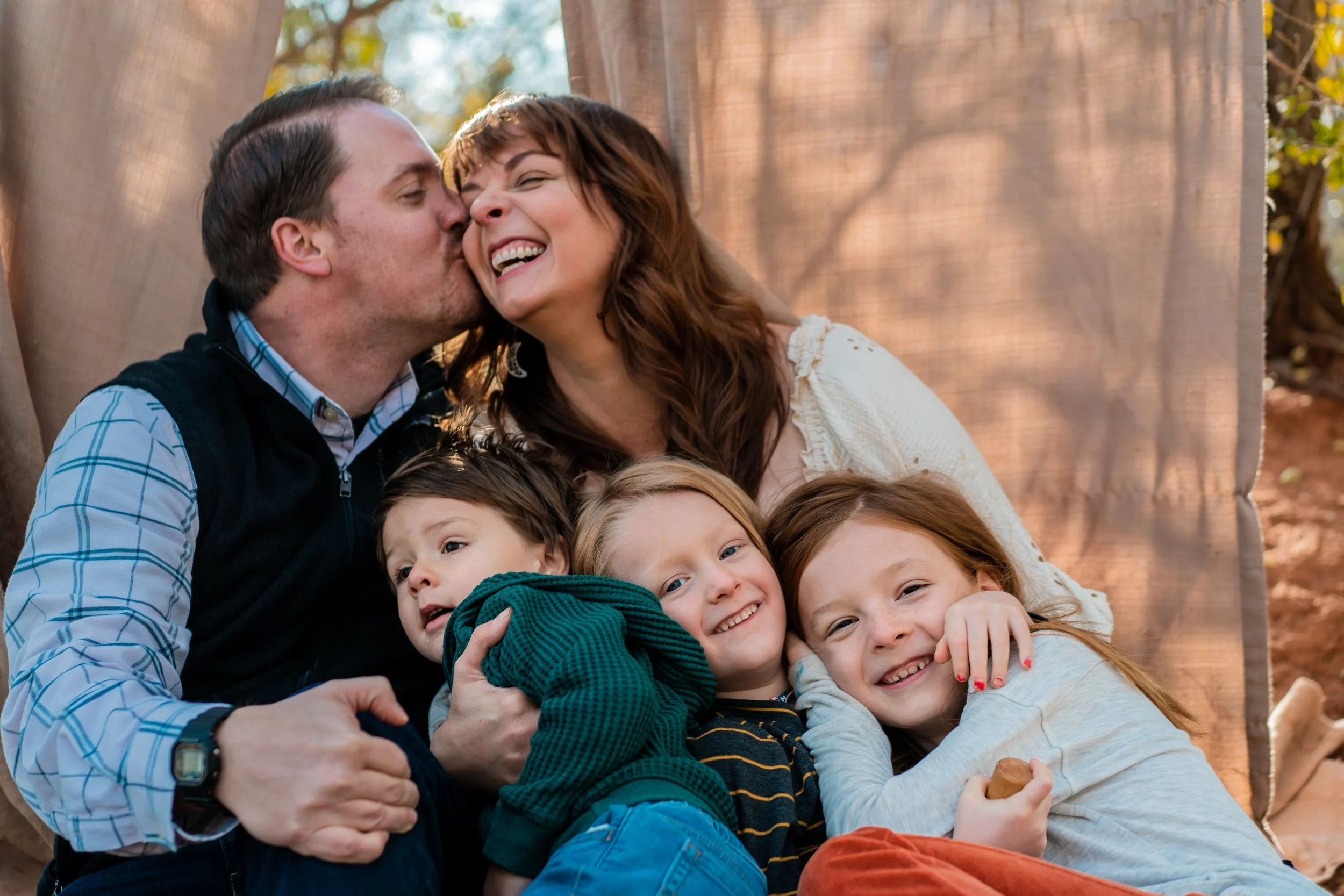 Family of five outdoors, with a man kissing a woman on the cheek while the children hug and smile.