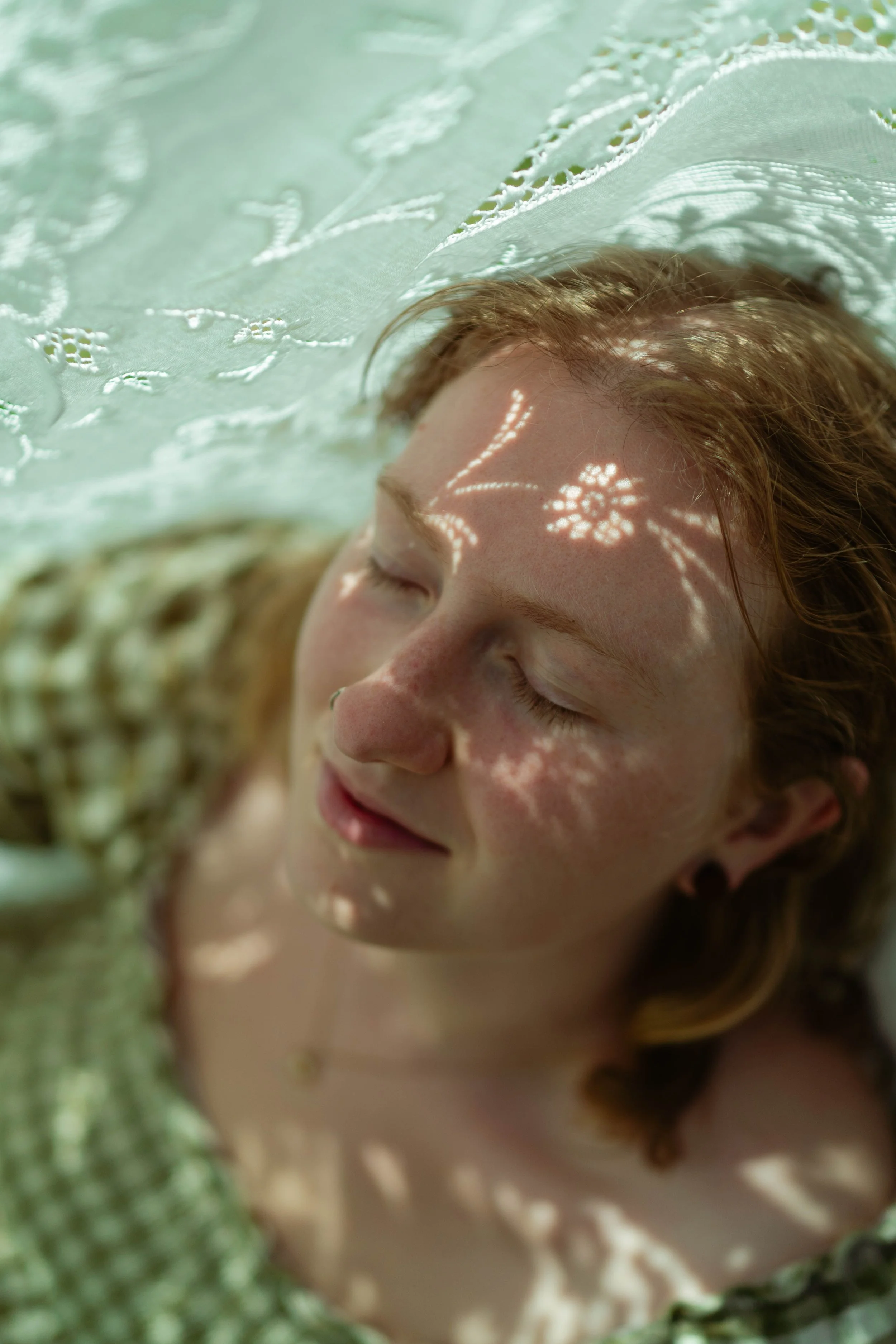 Close-up of a young woman's face with closed eyes, lying on a lace fabric that filters sunlight, creating floral shadow patterns on her face.